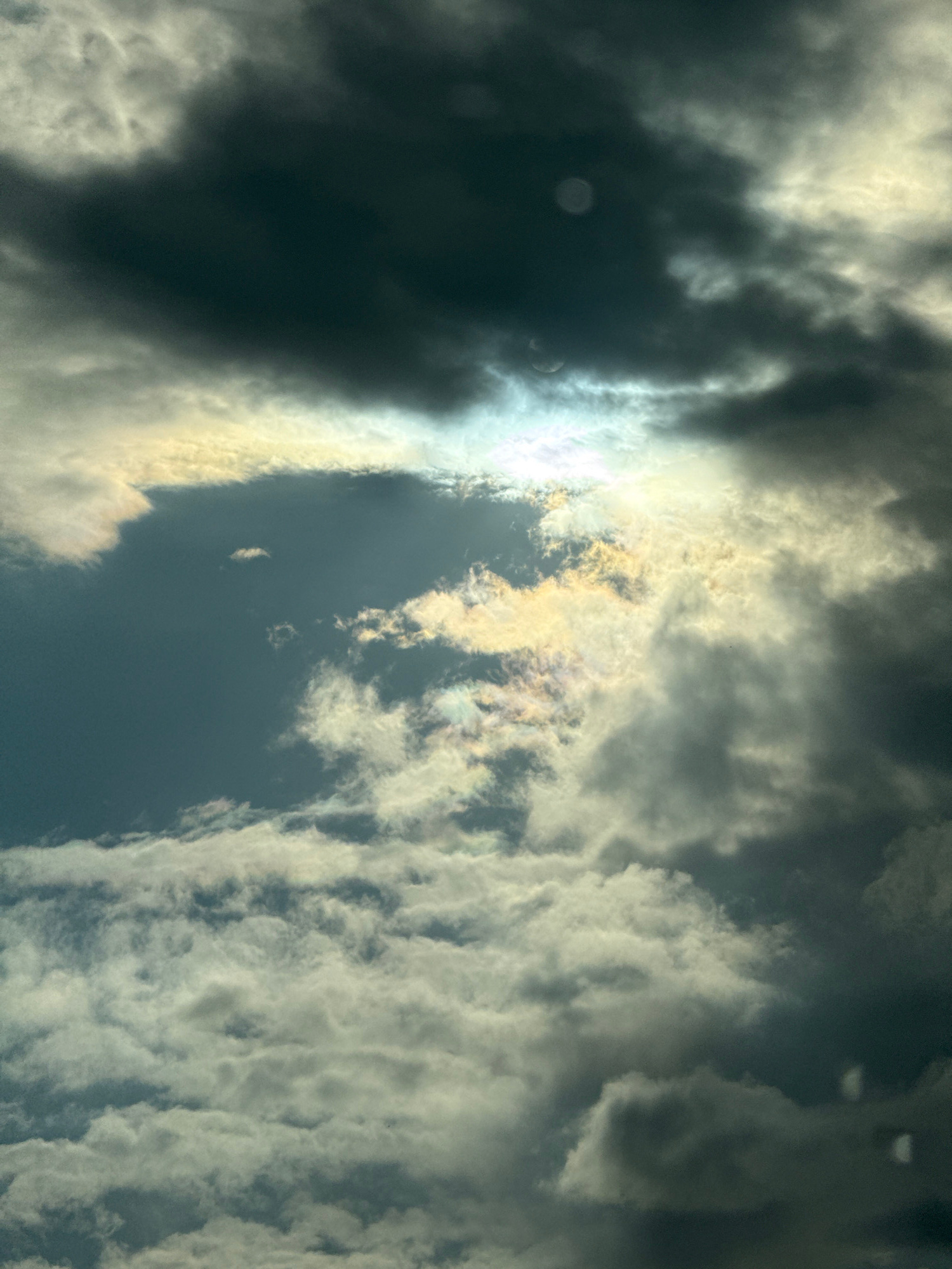 The image shows a dramatic sky filled with dark, heavy clouds interspersed with areas of bright, luminous light. The sunlight breaks through in a central portion, creating a bright, almost iridescent effect with streaks of yellow, white, and hints of pink and blue. The clouds are dense and appear to be moving, with some parts obscuring the light more than others. The overall impression is one of impending weather, perhaps a storm clearing or approaching. The scene is devoid of any identifiable structures, human figures, or specific landmarks that would place it within the city of Krasnogorsk, Russia, or any other specific urban environment. It is a purely atmospheric landscape.