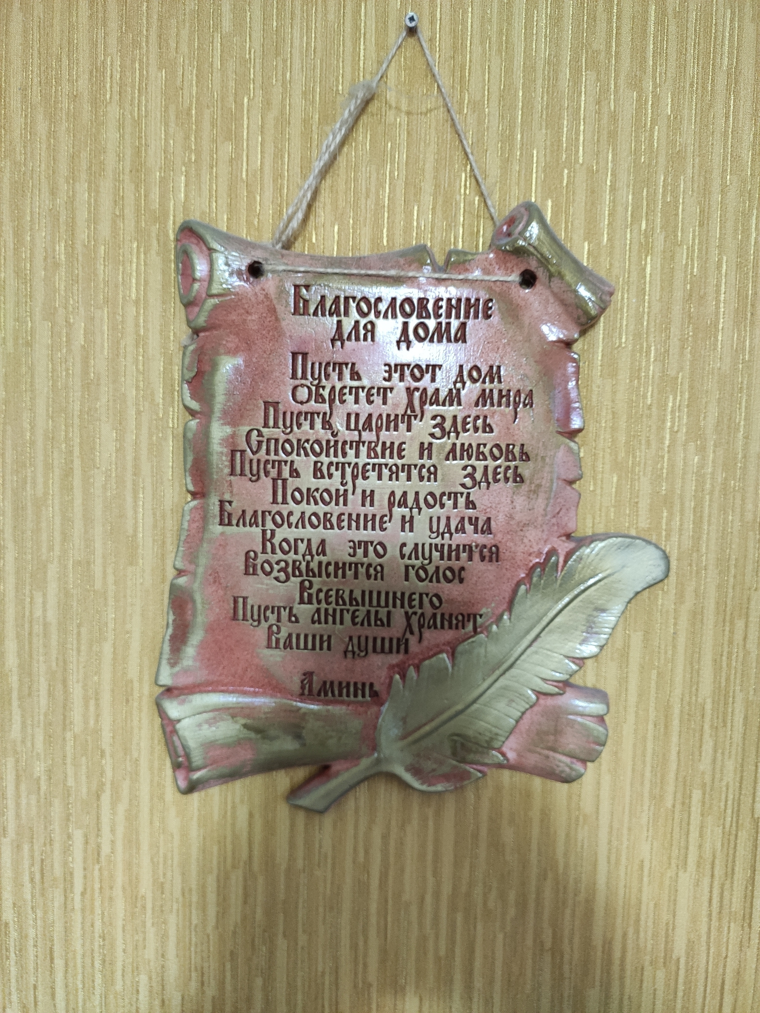 A decorative plaque made of what appears to be ceramic or plaster hangs from a string against a textured, yellowish wall. The plaque is shaped like a scroll and has a pinkish-red and silver color scheme with embossed text. The text is in Russian and is arranged in several lines. The top line reads "БЛАГОСЛОВЕНИЕ ДЛЯ ДОМА" which translates to "BLESSING FOR THE HOME". Below this are several more lines of text, which appear to be a prayer or blessing. The plaque also features an embossed image of a feather on the lower right side. The overall impression is of a decorative item intended for home display. There are no people or other discernible objects in the scene.