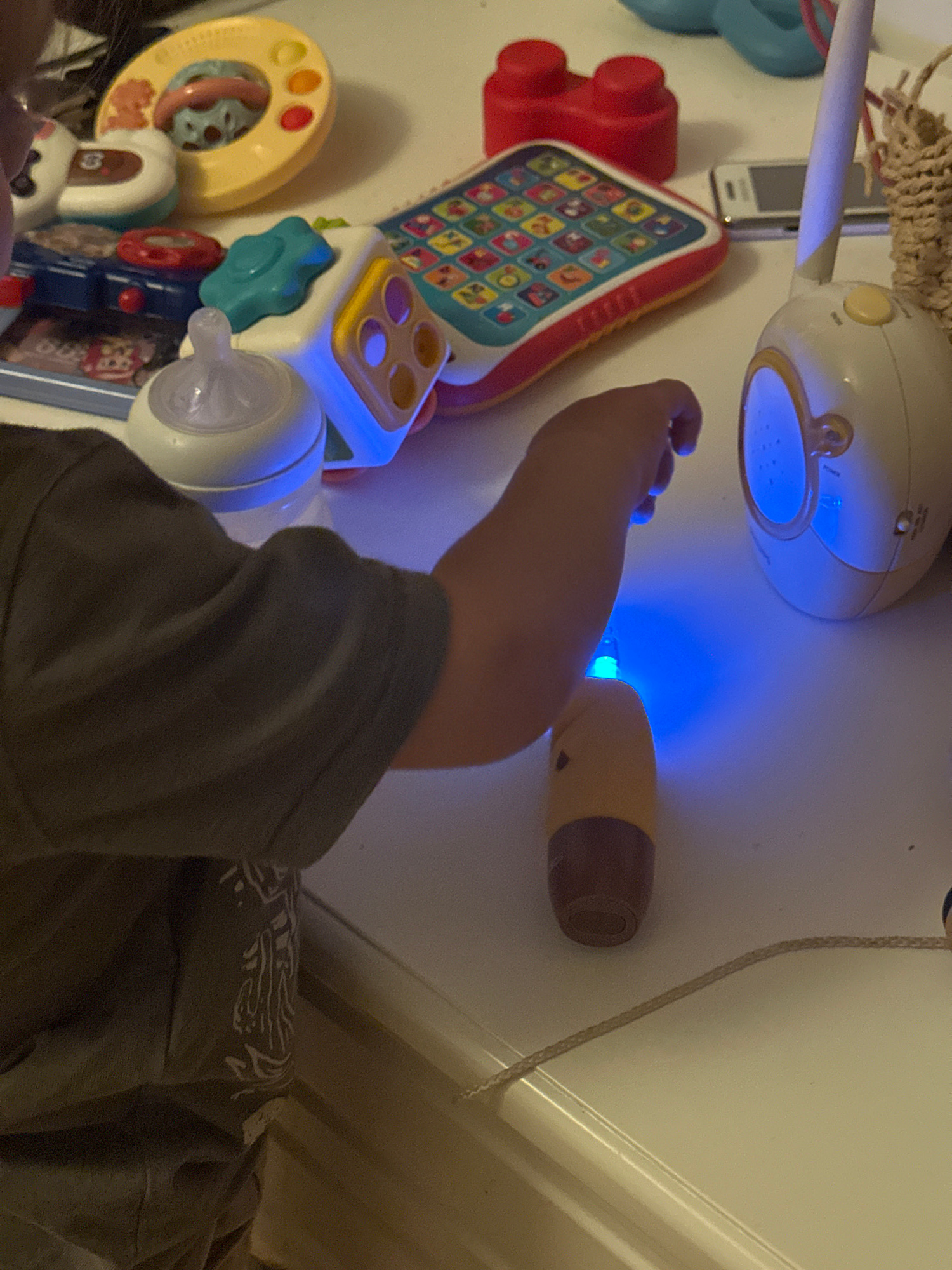 The image depicts a close-up view of a child's hands interacting with various toys on a white surface, likely a table or dresser. Several toys are visible, including a baby bottle with a blue light emanating from its base, a colorful electronic learning toy with buttons, a yellow ring-shaped toy, and red building blocks. A white baby monitor unit with a yellow button is also present. The child is wearing a dark t-shirt with a white graphic design on the sleeve. The lighting in the scene is dim, with the primary light source appearing to be the blue glow from the bottle. The image does not contain any identifiable landmarks or context that would allow for its location to be determined in Sofia, Bulgaria, or any other city.