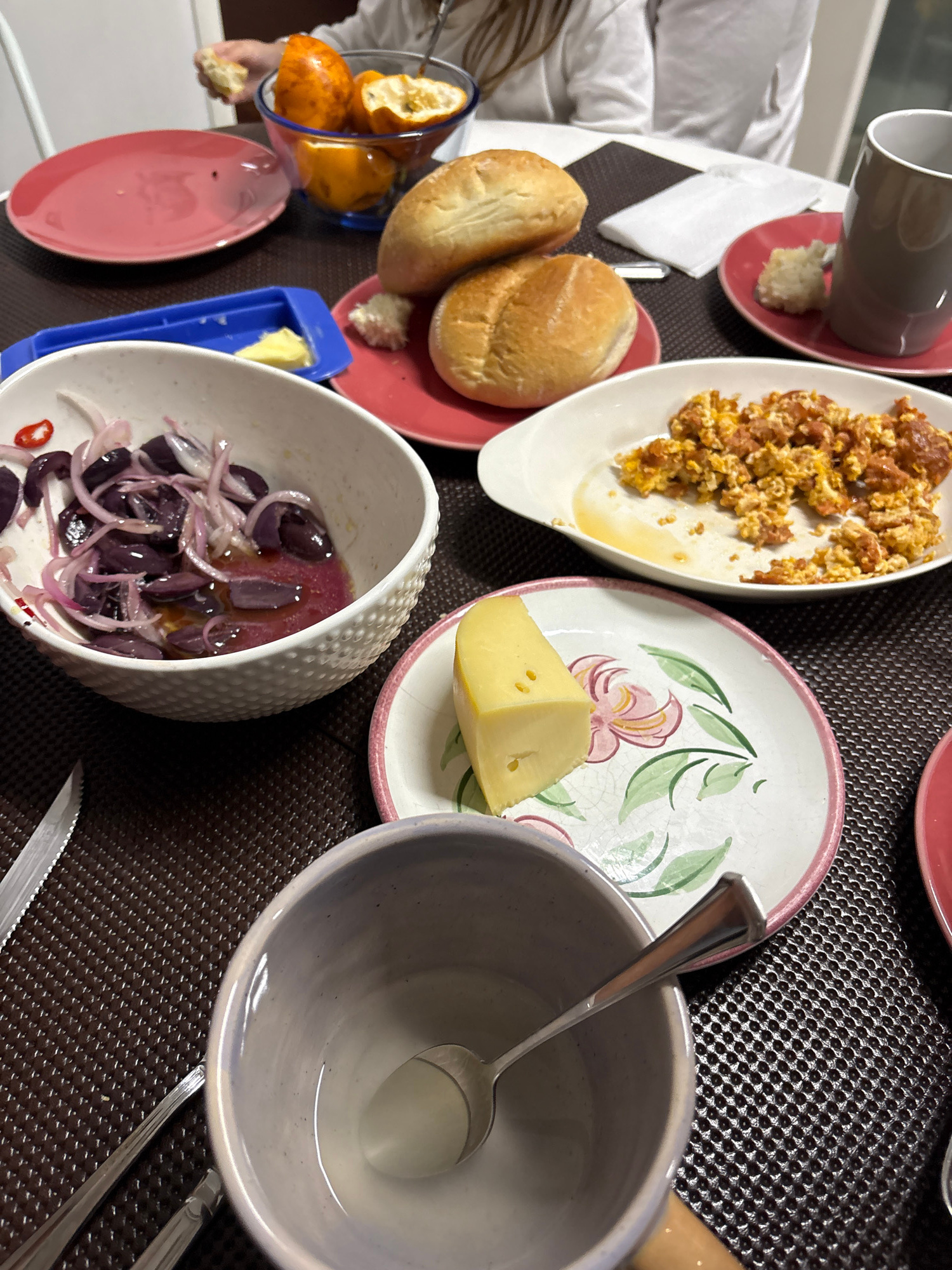 The image shows a table set for a meal, likely a breakfast or brunch, with several dishes and plates. In the foreground, a light purple bowl with a spoon rests on a textured brown placemat. To the left, a knife with a silver blade and a black handle is also on the placemat. A white bowl containing sliced purple olives and red onion, dressed in a vinaigrette, sits to the left of a pink plate holding a wedge of yellow cheese on a decorative plate with floral patterns. Behind the cheese plate, a white oval dish holds scrambled eggs mixed with what appears to be chorizo. Two round bread rolls are stacked on a pink plate to the left of a grey mug and a small pile of white rice or couscous. In the background, a person's hands are visible, holding a piece of bread. A clear blue bowl filled with sliced oranges or tangerines is also present on the table. The overall setting suggests a domestic dining environment. The image does not provide any specific visual cues that would allow for the localization to San Isidro, Peru.