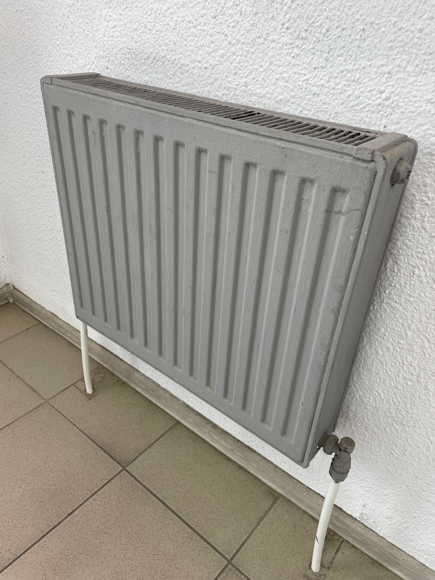 An elevated, slightly angled medium shot captures a gray radiator against a textured white wall. The radiator, designed with vertical fins, is supported by two white legs. To the lower right, a white pipe with a gray valve connects to the radiator. The floor is tiled with square, tan tiles. The scene is devoid of any people or any other discernible objects, creating a stark and minimalist composition.