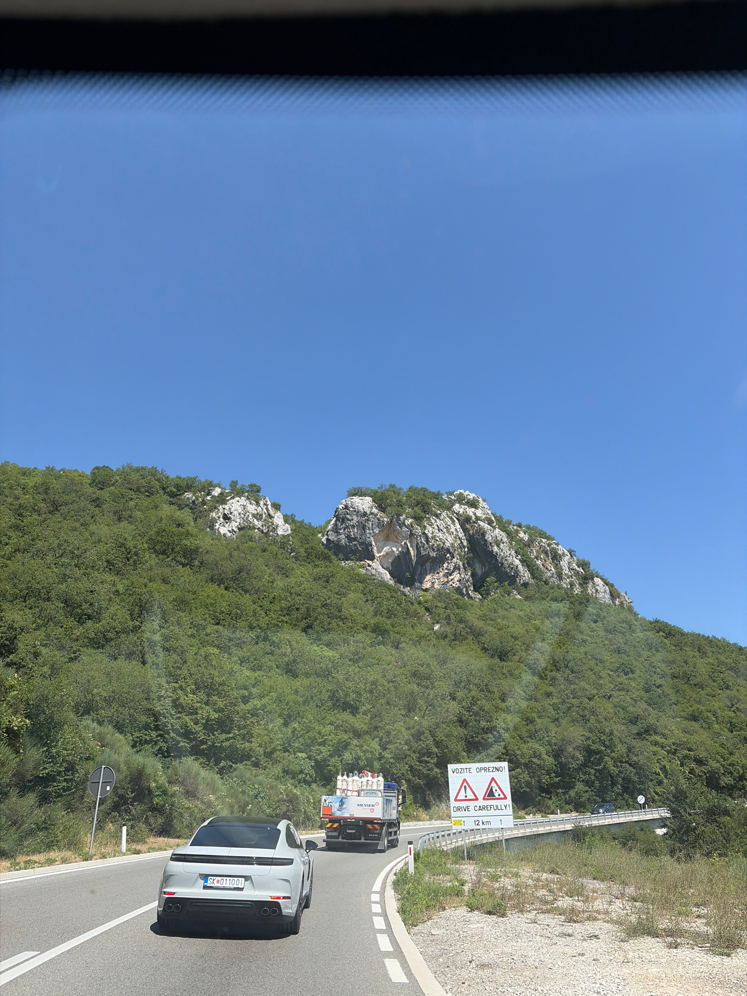 In Lapčići, Montenegro, a white Porsche Taycan, license plate SK-011001, travels on a paved road alongside a steep, tree-covered mountainside under a clear blue sky. Ahead of the Porsche, a truck loaded with white cylindrical containers, branded "Messer," proceeds along the same road. To the right, a white sign with red triangles, depicting falling rocks and a steep incline, displays the text "VOZITE OPREZNO! DRIVE CAREFULLY!" with "12 km" below. The road curves to the right, crossing a concrete bridge where another vehicle is visible in the distance. The environment is characterized by lush green vegetation covering the rocky mountainous terrain.