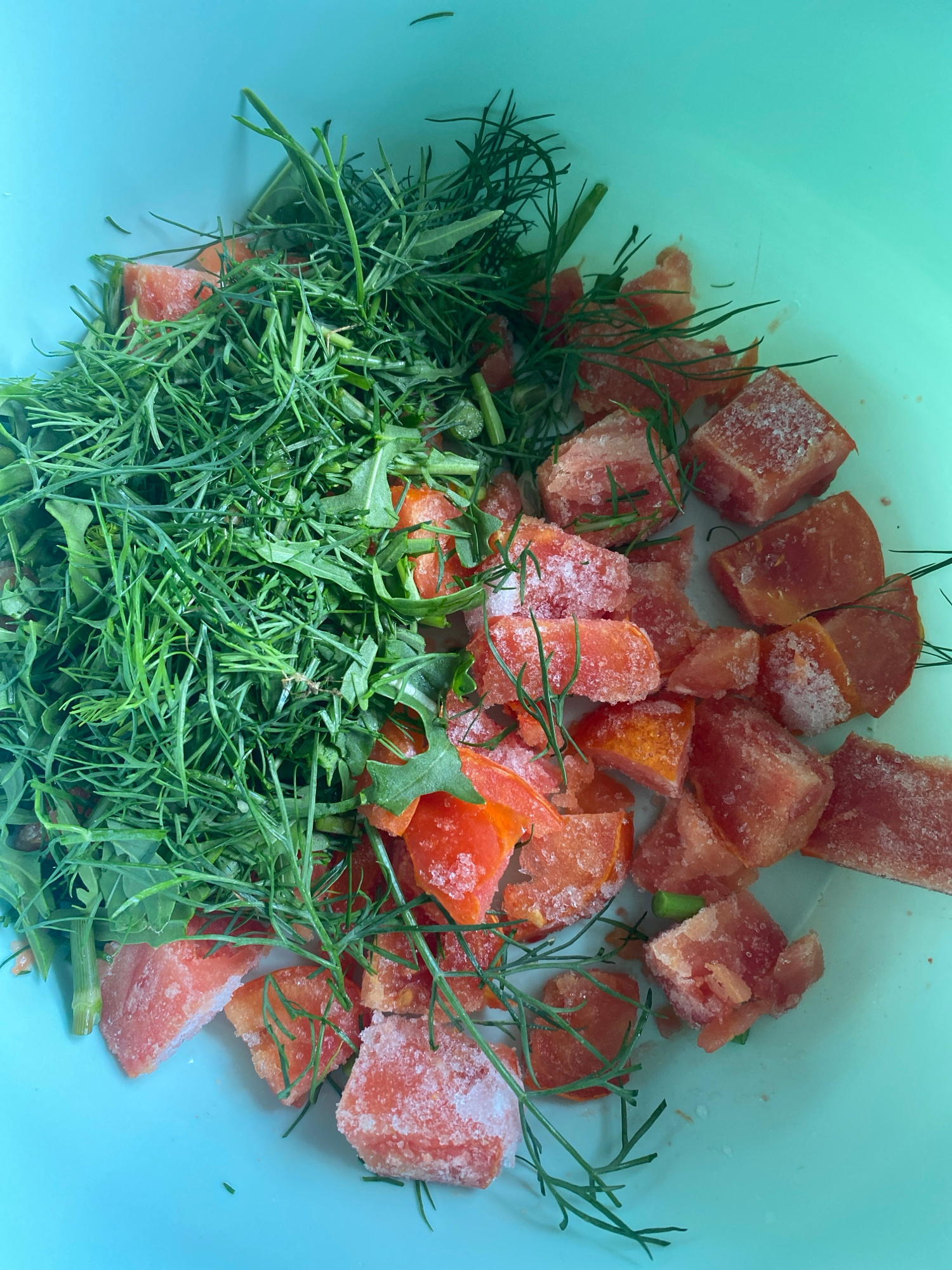 The image displays a top-down view of a light blue bowl filled with chopped red fruit, possibly tomatoes, and green herbs. The fruit pieces are mostly cubed and appear to be frozen, with a white frosty coating visible on some of them. The green herbs are finely chopped and interspersed among the fruit. The bowl is sitting on a flat, light blue surface, which could be a countertop or a table. There are no people visible in the image. The scene is likely indoors, given the lighting and the smooth, artificial surface of the bowl. The image does not contain any information or context that would allow for the determination of its location within the city of Krupki, Belarus.
