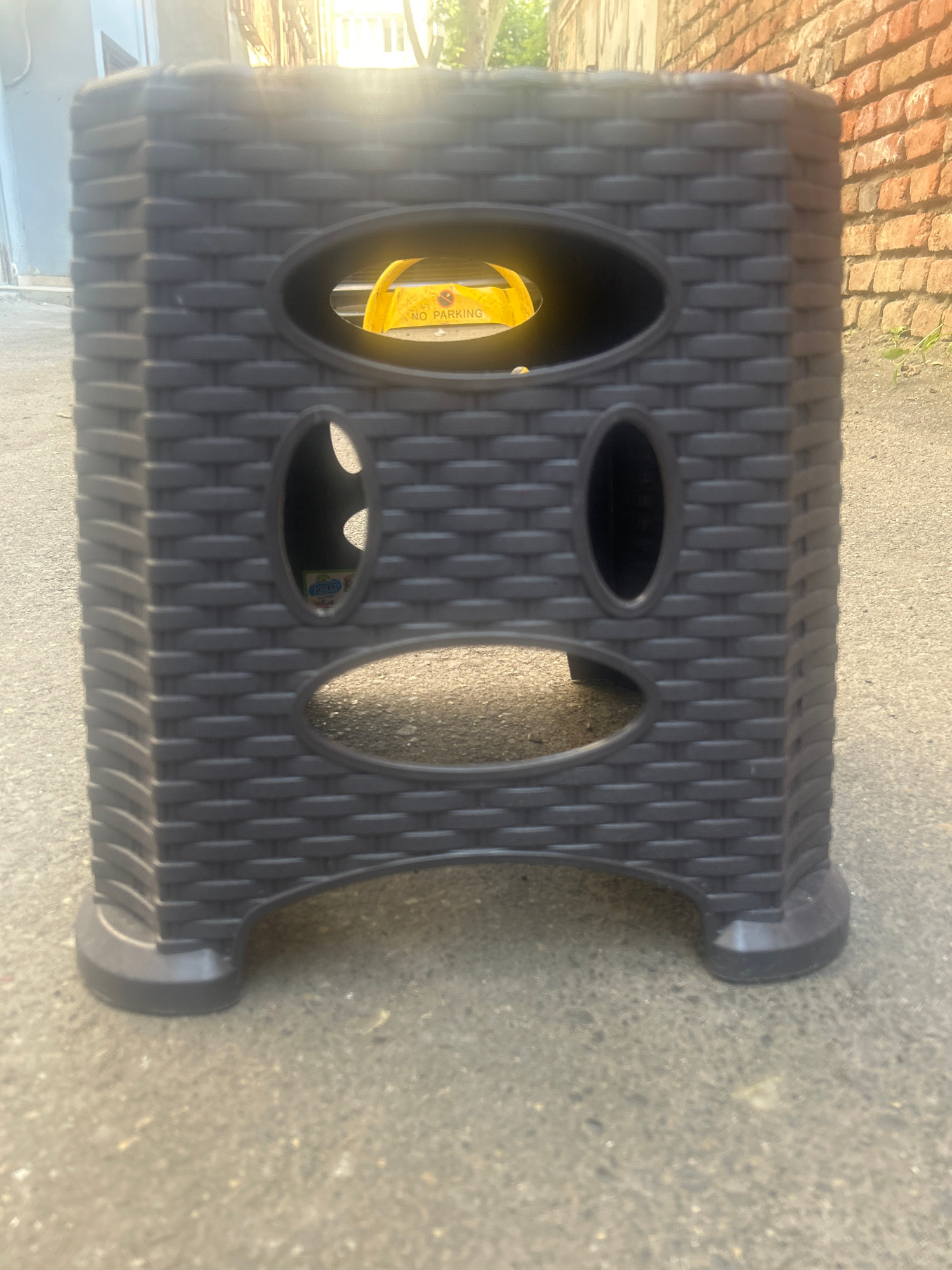 The image shows a dark grey, plastic, rectangular stool with an oval-shaped opening on each of its four sides. The stool has a woven texture pattern. Inside one of the openings, a yellow strap with black text is visible. The stool is positioned on a grey asphalt surface, likely outdoors, with a brick wall visible in the background on the left. The lighting suggests it is daytime, possibly late afternoon, with sunlight creating shadows. The scene appears to be a street or sidewalk in Tbilisi, Georgia. No people are visible in the image. The visible text on the yellow strap reads "NO PARKING".