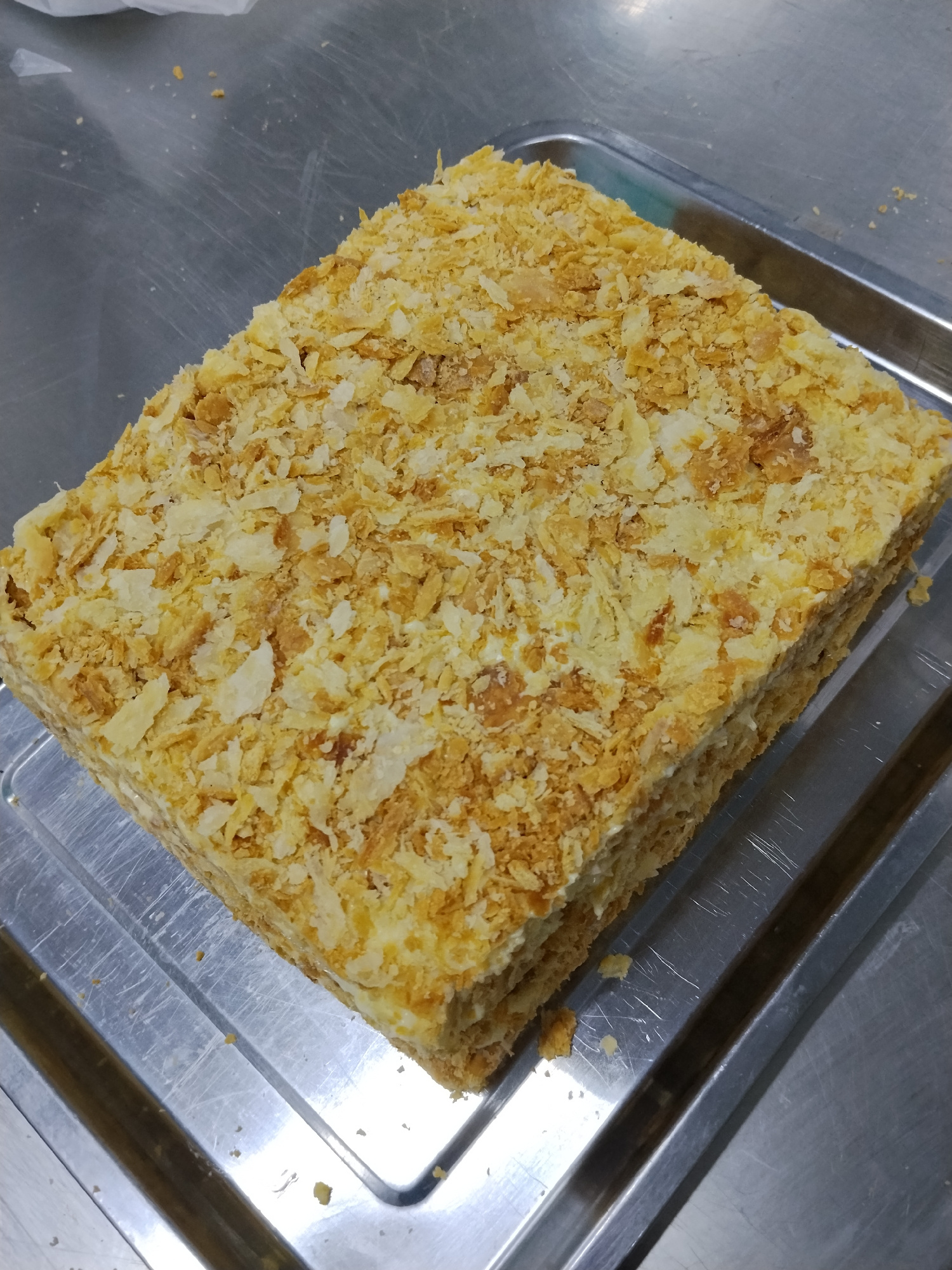 A rectangular cake covered in crumbled pastry is presented on a silver tray, which is placed on a metal surface. The cake has multiple layers of pastry and cream, with the top and sides generously coated in the crumbled topping. The overall impression is that of a homemade or freshly prepared dessert. The location is not determinable from the image.