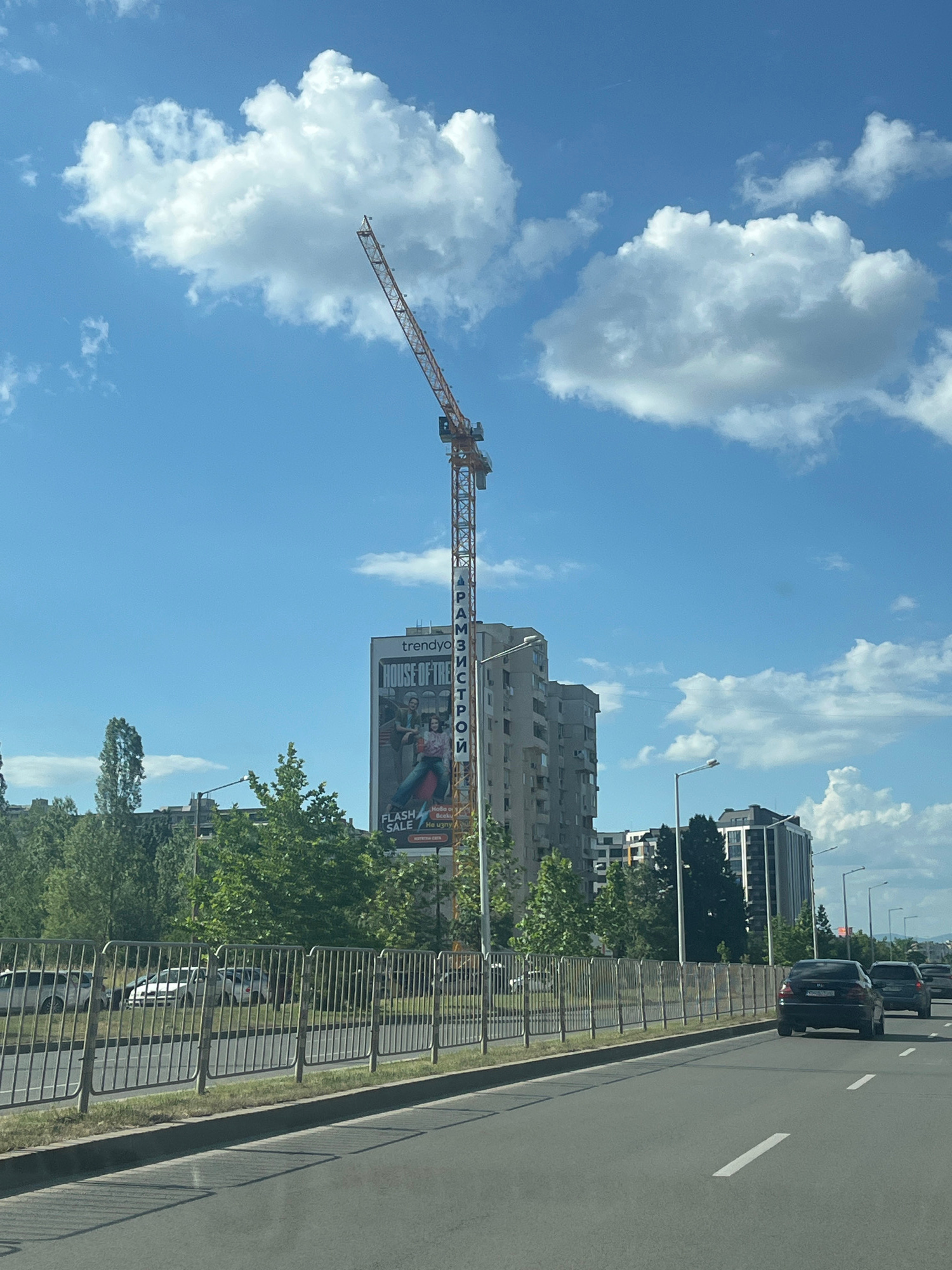 The image shows a construction crane towering over a building in Sofia, Bulgaria. The building has a large advertisement on its side for "trendyyo HOUSE OF THE TRENDYLO" with text that appears to be in Cyrillic script. The advertisement also mentions "FLASH SALE". The scene is viewed from a road with cars driving by. The sky is blue with white clouds. The overall impression is of a modern urban environment with ongoing construction and commercial activity.