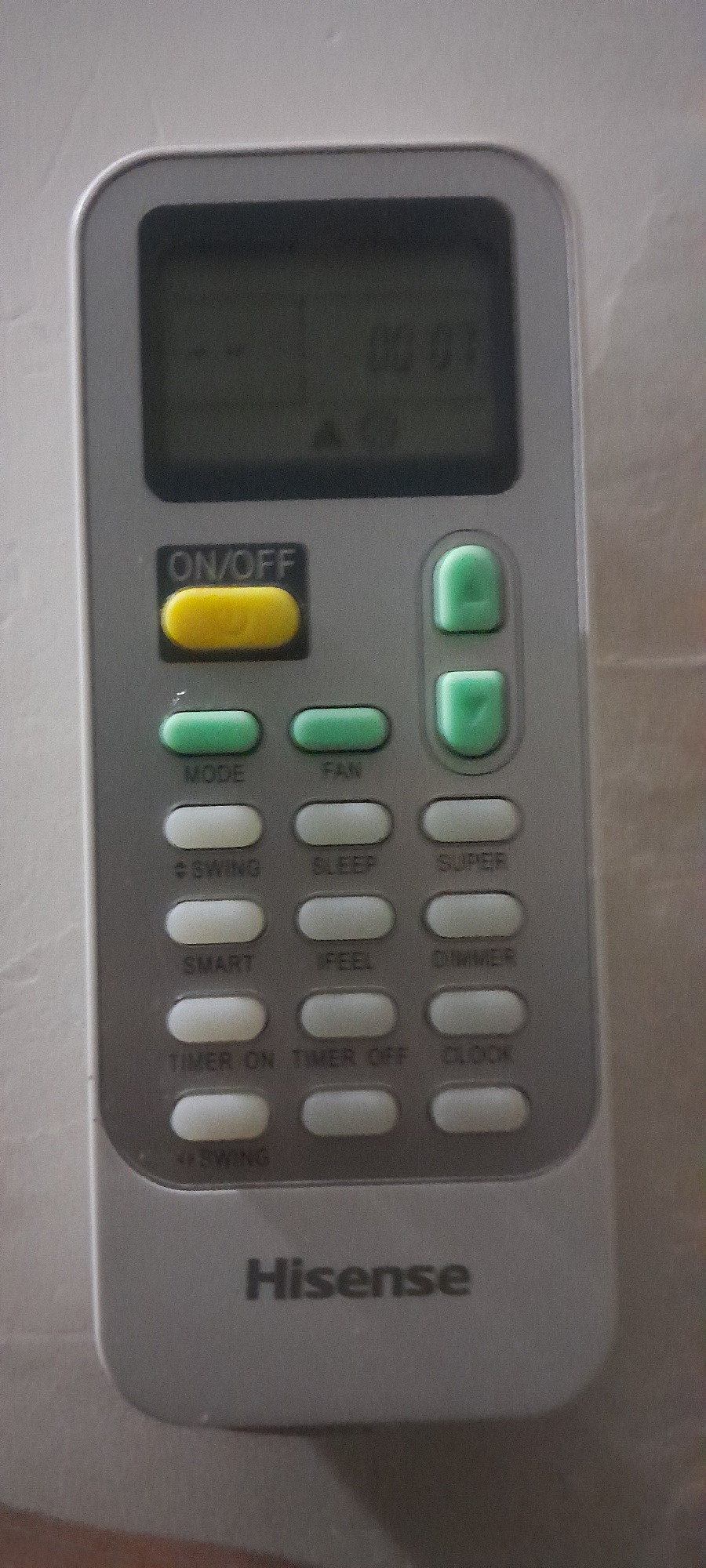 The image shows a Hisense air conditioner remote control on a light gray, textured surface, possibly a wall or countertop. The remote is silver with black and colored buttons. The top of the remote has a digital display showing numbers and symbols, with "00:07" visible. Below the display, there's a black button labeled "ON/OFF" with a yellow oval button underneath. To the right of the ON/OFF button are two green buttons, one above the other. The rest of the remote is covered in rows of gray and white oval buttons with various labels such as "MODE," "FAN," "SWING," "SLEEP," "SUPER," "SMART," "I FEEL," "DIMMER," "TIMER ON," "TIMER OFF," and "CLOCK." The brand name "Hisense" is printed in black text on the lower part of the remote.