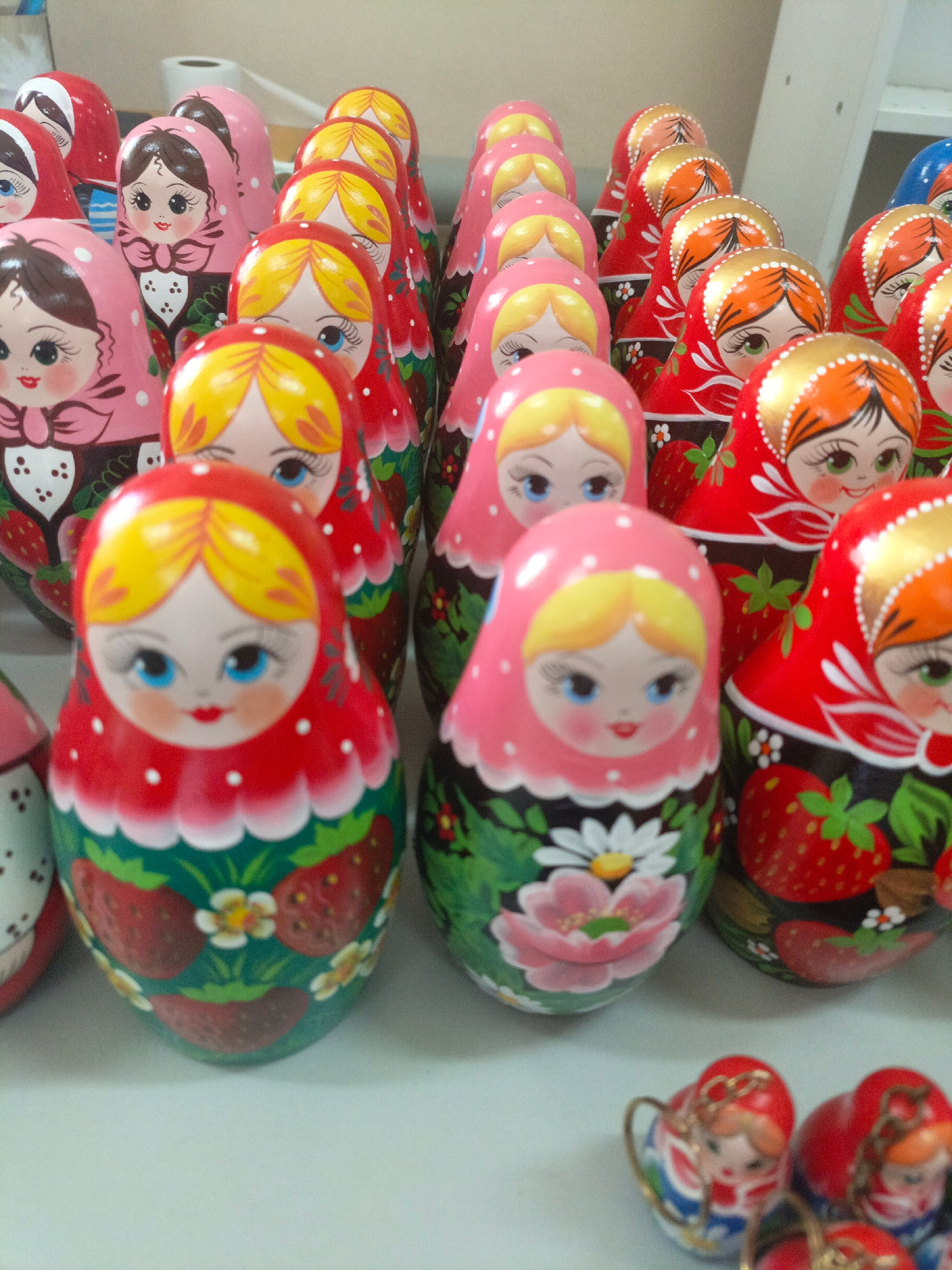 In the foreground, several Russian nesting dolls are displayed on a light-colored surface. The dolls vary in size and design, with some featuring floral patterns and others depicting traditional Russian folk art. The dolls are arranged in rows, with the largest ones in the front and smaller ones behind. The dolls are made of wood and painted with bright colors, including red, pink, yellow, and green. The surface they are on appears to be a table or shelf. In the background, a white shelf unit is visible, suggesting the dolls are in a store or workshop. There is no indication of a specific location in the city of Brest, Belarus.