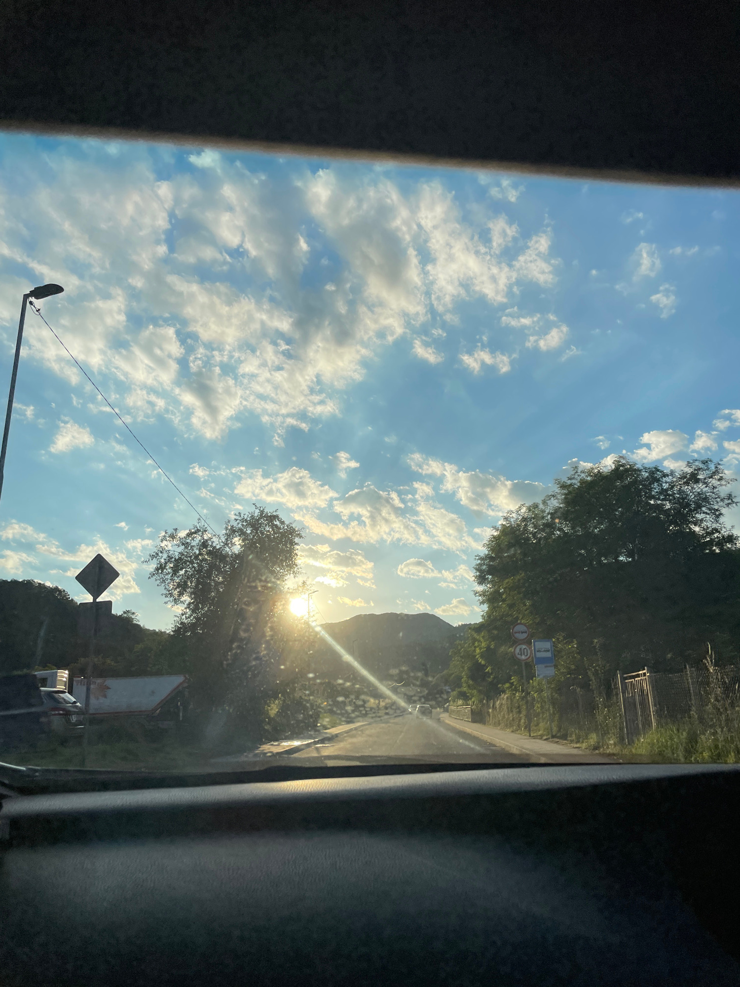 The image captures a view from inside a vehicle looking through the windshield. The scene depicts a road bathed in the warm light of a setting sun, which is positioned behind a hill in the distance. Rays of sunlight pierce through a sky filled with scattered white clouds, creating a dramatic effect.
On the left side of the road, a street lamp stands tall, and a diamond-shaped warning sign is visible. Further along the road, a large tree partially obstructs the view, with the sun's glare creating a lens flare effect. To the right of the road, there is a metal fence and a bus stop sign with a speed limit indicator of 40. A few vehicles are present, including a parked car and a trailer on the left side. The overall environment suggests a rural or suburban setting. The presence of the bus stop and road infrastructure points to a frequented route within the city of Zlatograd, Bulgaria.