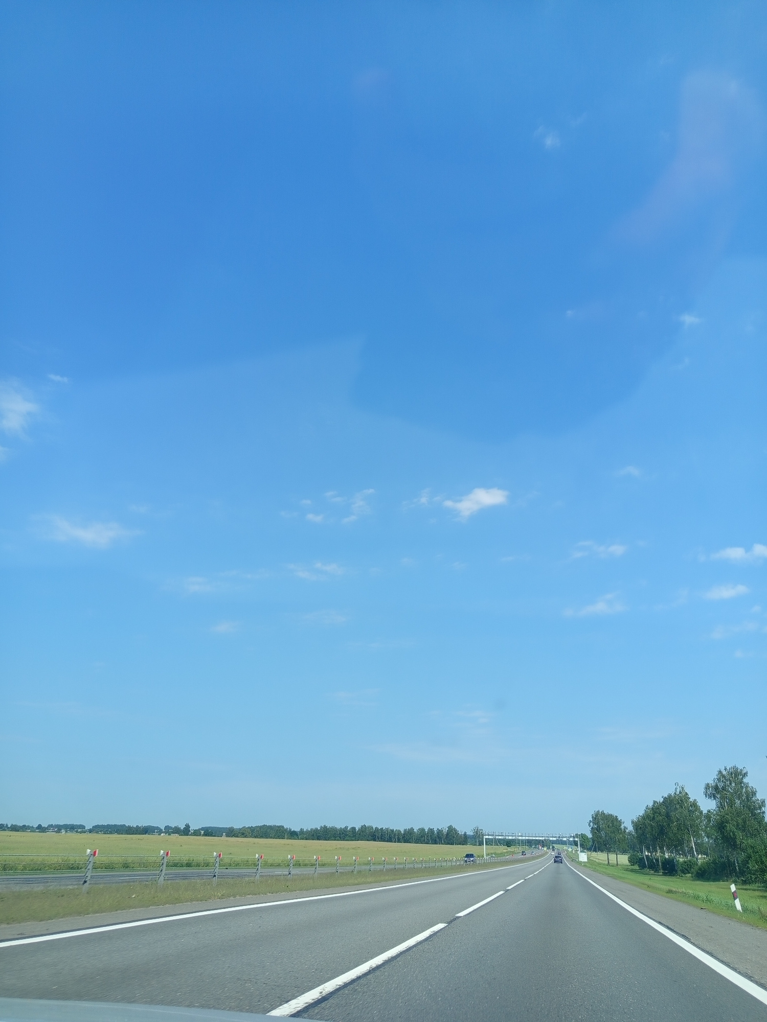 The image depicts a highway scene on a clear, sunny day in the vicinity of Валевачскі сельскі Савет, Belarus. The perspective is from a vehicle traveling on a multi-lane asphalt road. The road is bordered by white lines and guardrails with red and white markers. To the left of the road, a green field extends to the horizon, dotted with sparse trees and structures. To the right, a grassy area is followed by a line of trees. In the distance, a highway overpass can be seen, with a few vehicles on the road. The sky is a vibrant blue with scattered white clouds. There are no people visible in the image.