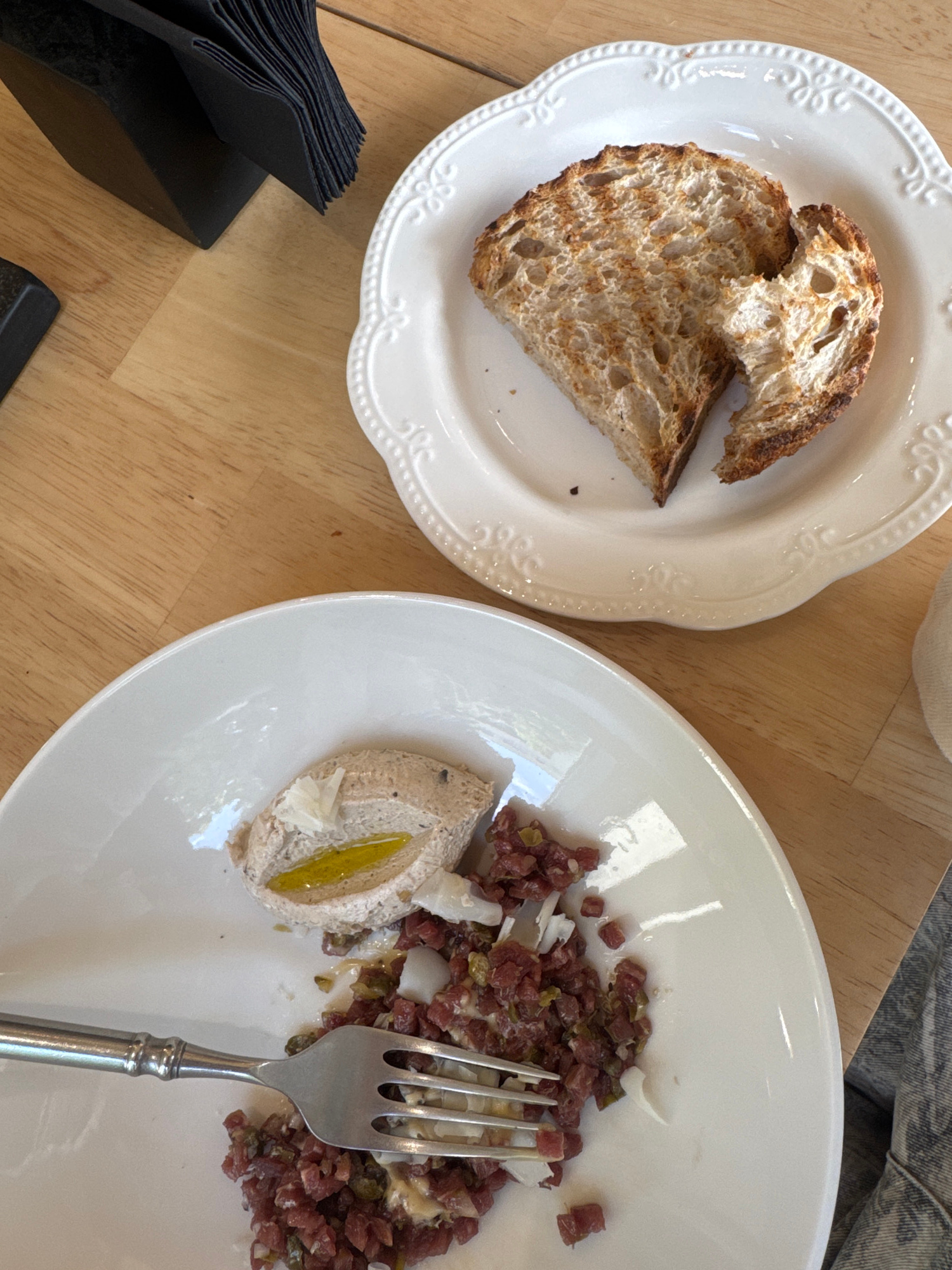 An overhead, close-up shot captures a meal on a wooden table. In the foreground, a white plate holds a serving of what appears to be tartare, a creamy spread topped with a sliver of yellow pepper, and a scattering of small, diced ingredients. A silver fork rests on the plate, partially obscuring the tartare. In the background, another white plate contains two slices of toasted bread, one with a bite taken out of it. To the left, a black napkin holder is filled with black napkins. The scene is set in Warsaw, Poland, at a dining establishment. There are no people visible in the image.