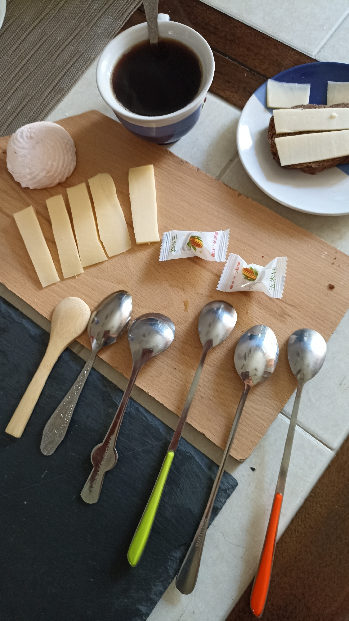 The image displays a close-up, top-down view of a table setting with breakfast items. A white mug with blue trim, filled with dark coffee, sits on a textured placemat. A metal spoon with a patterned handle is in the mug. To the left of the mug is a light pink, swirled meringue or marshmallow.
In the foreground, a wooden cutting board holds several rectangular slices of pale yellow cheese. Next to the cheese are two small, individually wrapped candies with Asian characters on them.
Below the cutting board, a dark gray slate placemat is visible, on which five spoons are arranged. One is made of wood, and the other four are metal. The metal spoons have different colored handles: one is green, one is orange, and the other two appear to be silver. Some of the metal spoons have visible decorative etching on their handles.
To the right of the coffee mug, on a blue and white plate, is a slice of dark bread topped with three rectangular slices of the same pale yellow cheese. The table surface appears to be a tiled floor or tabletop with diagonal lines, possibly wood grain or tile grout. The background shows glimpses of other table surfaces, one a dark brown wood and another a lighter, textured material. There is no indication of the city of Kapyĺ, Belarus, or any specific geographical location within the image itself.