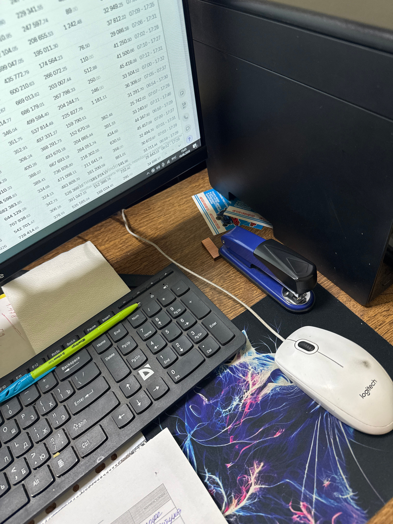 The image shows a desk with a computer keyboard, a white Logitech mouse, a blue stapler, and a tablet displaying a spreadsheet. A green pen rests on the keyboard. The scene is set on a wooden desk, with a patterned mousepad beneath the mouse and keyboard. The tablet screen displays a financial or data-heavy spreadsheet with numerous columns and rows of numbers, suggesting a work or study environment. The lighting suggests it is daytime. The text visible on the tablet screen and on a small card near the stapler appears to be in Russian. The card has text that translates to "Construction of drains" and "Access to yards" along with phone numbers. The tablet screen also shows a date "19.06.2026" and time "15:05". The overall impression is a personal workspace, likely used for tasks involving data analysis or record-keeping. There are no people visible in the image.