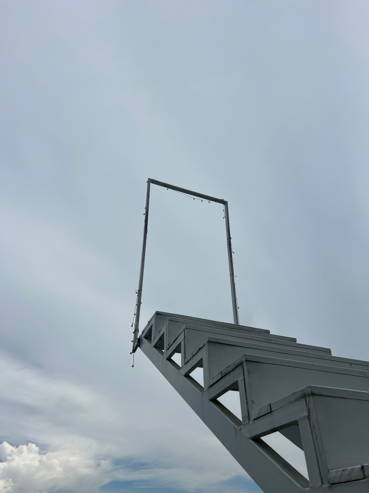 A low-angle, full shot captures a white, angular staircase extending diagonally upward into a cloudy, overcast sky. The staircase is constructed of metal beams with triangular cutouts, and the steps have a clean, modern design. At the top of the staircase, a thin, rectangular frame, also outlined in white, stands upright, adorned with a string of small, globe-shaped lights. The frame appears to be open, suggesting a passage or doorway. The sky is filled with a gradient of grey and white clouds, with a brighter expanse at the upper right. The overall impression is one of an abstract, minimalist structure set against a muted sky. The location of Baan Thurian, Thailand is not discernible from the visual elements of the image.