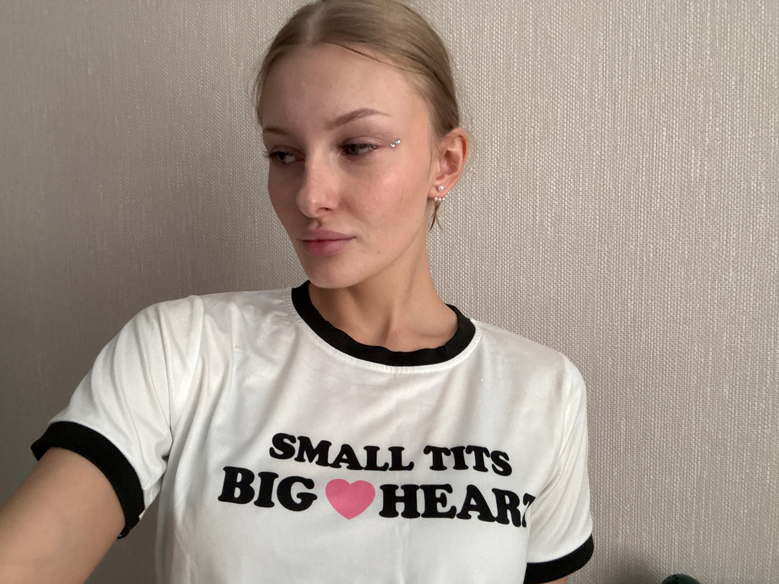 A young woman with blonde hair pulled back and light makeup is looking to the left. She is wearing a white t-shirt with black trim and the text "SMALL TITS BIG HEART" printed on it, with a pink heart symbol replacing the letter "O" in "HEART". She has a small silver stud above her right eyebrow and pearl earrings. The background is a textured light gray wall. The scene appears to be indoors, likely a domestic setting. There is no identifiable information about the city of Perm, Russia.