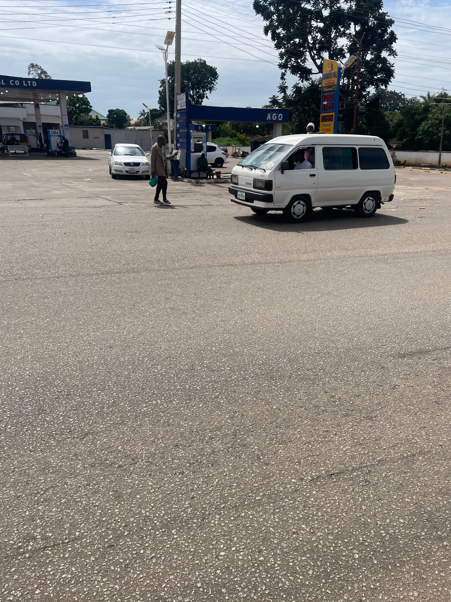 A white Toyota Hiace van drives on an asphalt road in front of a gas station in Jos, Nigeria. The gas station has a blue awning and a tall yellow sign displaying fuel prices. A person walks across the road in the foreground, holding a green object. Other vehicles and people are visible at the gas station. The sky is partly cloudy.