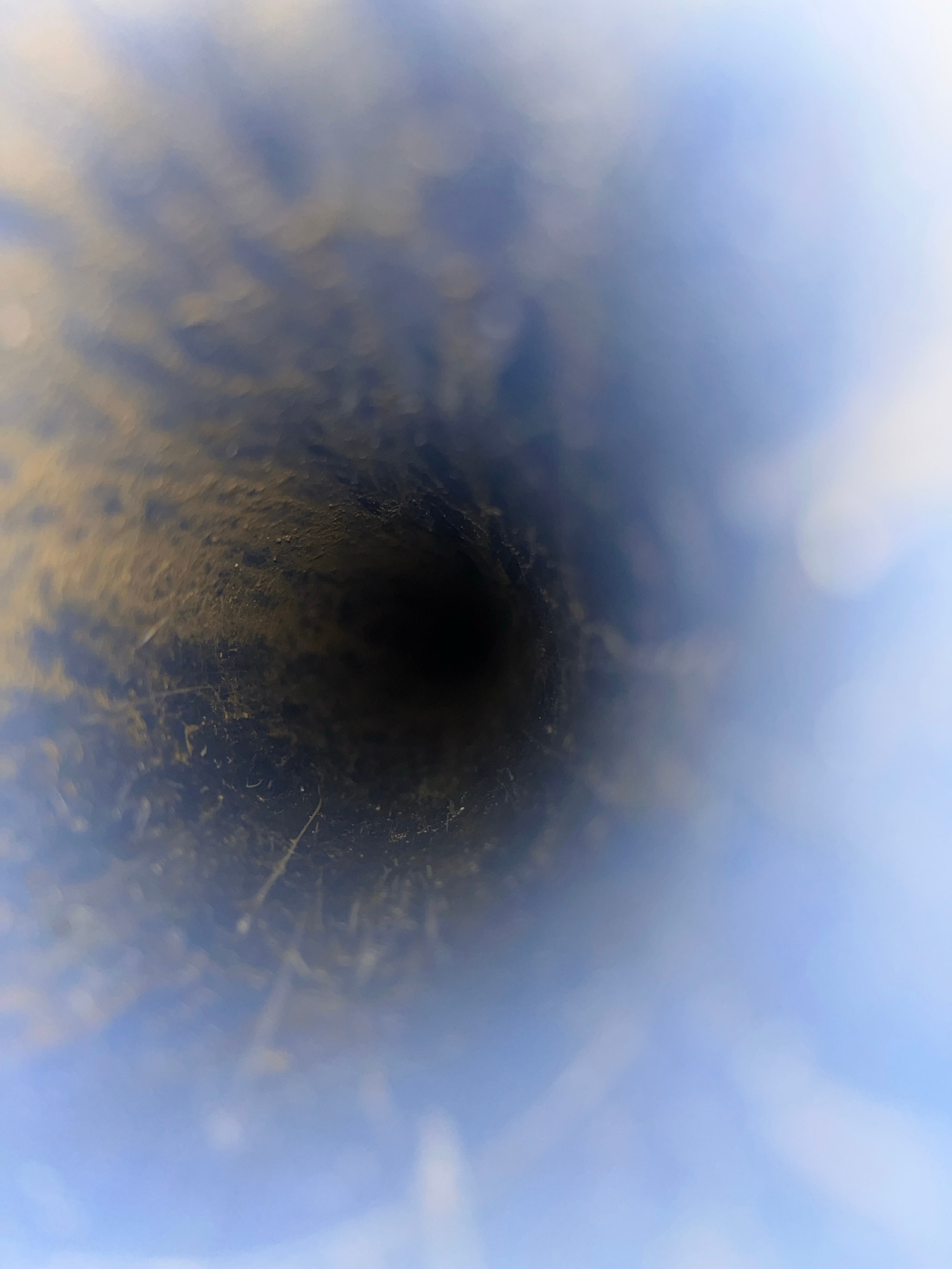 The image shows a narrow, dark tunnel-like opening with rough, textured walls. The opening recedes into darkness, creating a sense of depth. The edges of the tunnel are blurred and out of focus, with lighter, hazy blue and white areas suggesting light or possibly a sky. There appear to be small specks and debris on the inner surfaces of the tunnel. The overall impression is of looking into a hole or a pipe, possibly made of earth or a rough material. The lighting is dim within the tunnel and brighter and diffused on the exterior. There is no text visible in the image. The context provided of "Kota Kuala Simpang, Indonesia" suggests this could be a natural formation, a man-made structure like a well or a drainage pipe, or an entrance to an underground space in that region. No people, animals, or specific objects are discernible. The mood is ambiguous, potentially mysterious or claustrophobic due to the confined perspective and darkness.
