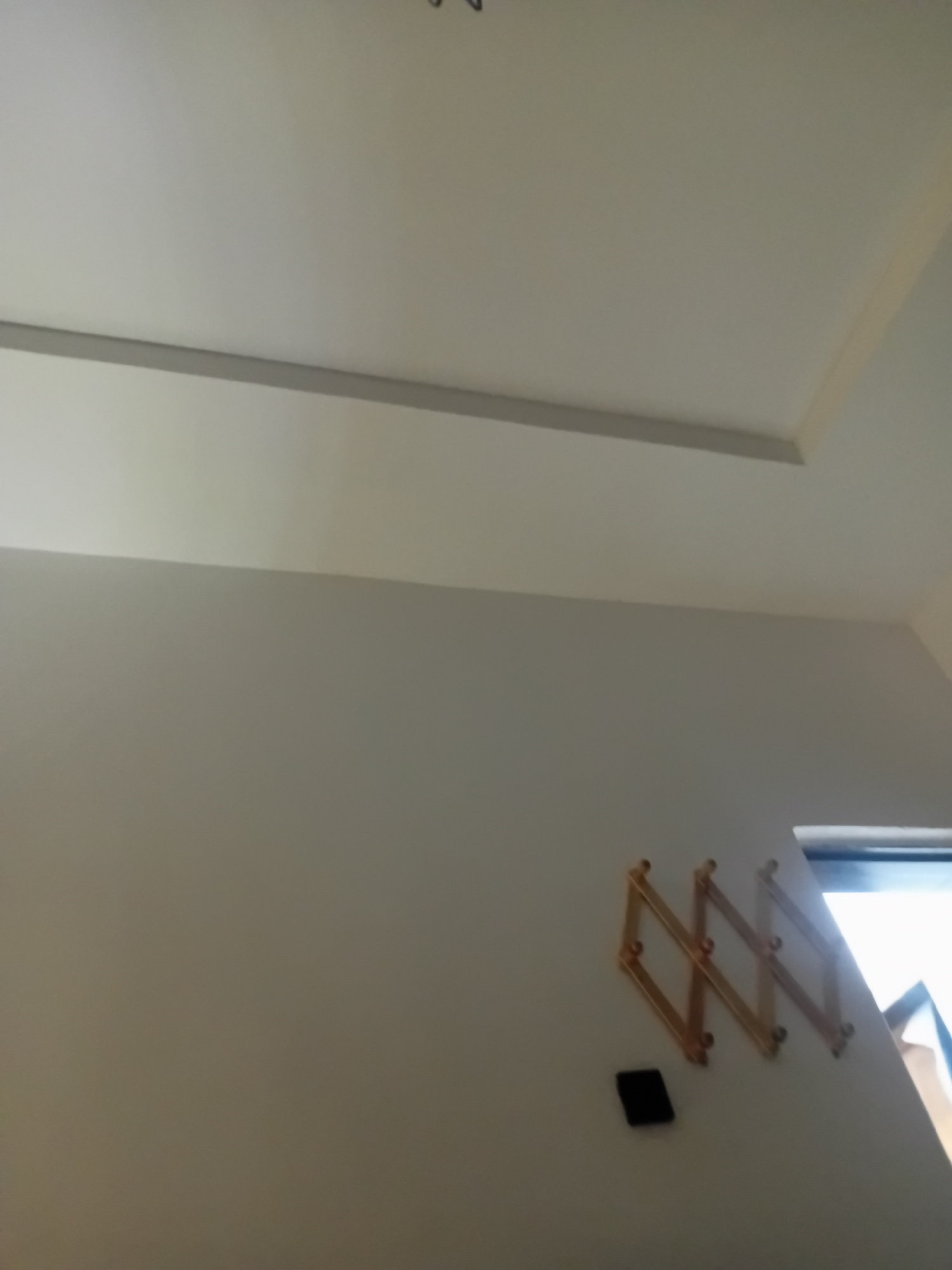 The image depicts an interior wall with a sectional ceiling and a window. Mounted on the wall is a collapsible wooden drying rack, resembling a series of interconnected diamonds. Below the drying rack, a small black object is attached to the wall. A portion of a window frame is visible to the right, with bright light emanating from outside. The walls and ceiling are painted in neutral, light colors. The provided image does not contain any information or visual cues that would allow for the determination of its location within the city of Kwang, Nigeria, or any other specific geographical area.
