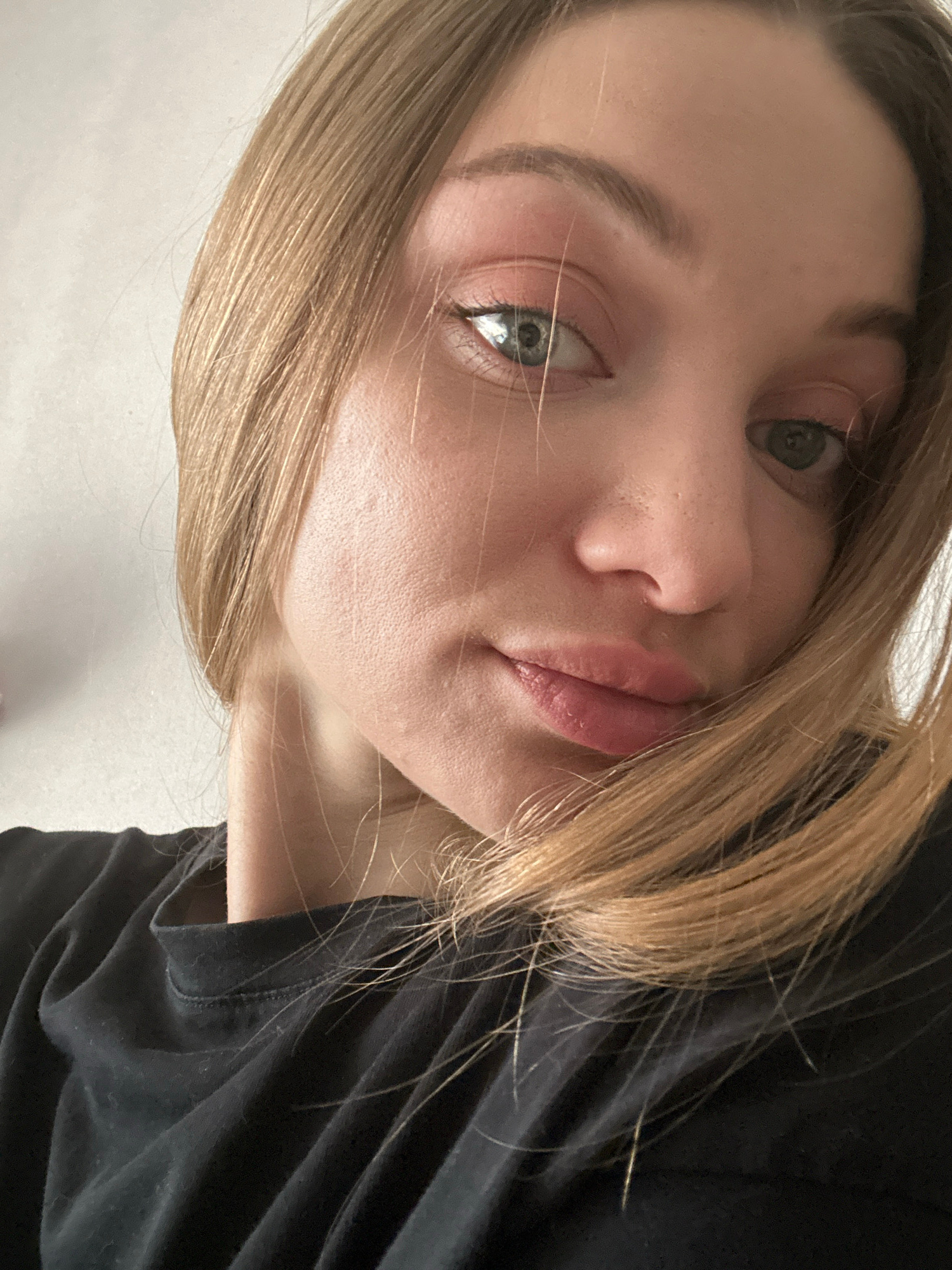 This is a close-up, angled selfie of a young woman with blonde hair and light-colored eyes. She is wearing a black shirt. Her face is tilted towards the camera, with her eyes looking slightly upwards and to the side. Her lips are parted, and she appears to be looking at the camera. The lighting suggests an indoor setting, possibly during the daytime, given the soft natural light on her skin. The background is a plain, light-colored surface, with some darker fabric visible on the right side of the frame, likely her clothing. There is no visible text in the image. The overall impression is a casual, informal portrait.