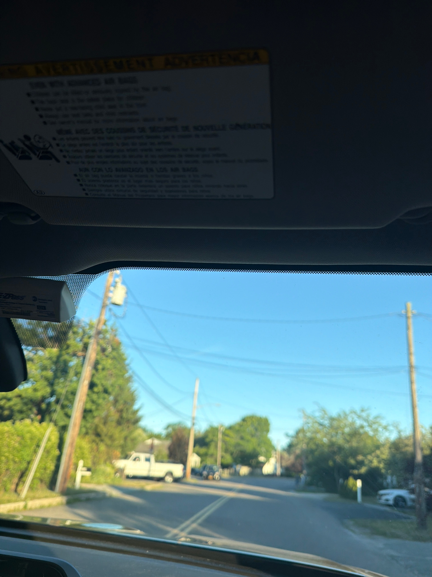 The image captures a view from inside a vehicle, looking out through the windshield. A sun visor, with warning labels in English, French, and Spanish, is visible at the top of the frame. A small electronic toll pass is attached to the windshield on the right side. The exterior scene shows a clear blue sky and a residential street lined with trees and power poles. A white pickup truck is parked on the side of the road, and a few other vehicles are present further down the street. The overall environment suggests a suburban or semi-rural setting, possibly in Riverhead, United States of America, given the context. There are no people or animals directly visible in the frame. The lighting indicates it is daytime, likely late afternoon or early evening due to the long shadows.