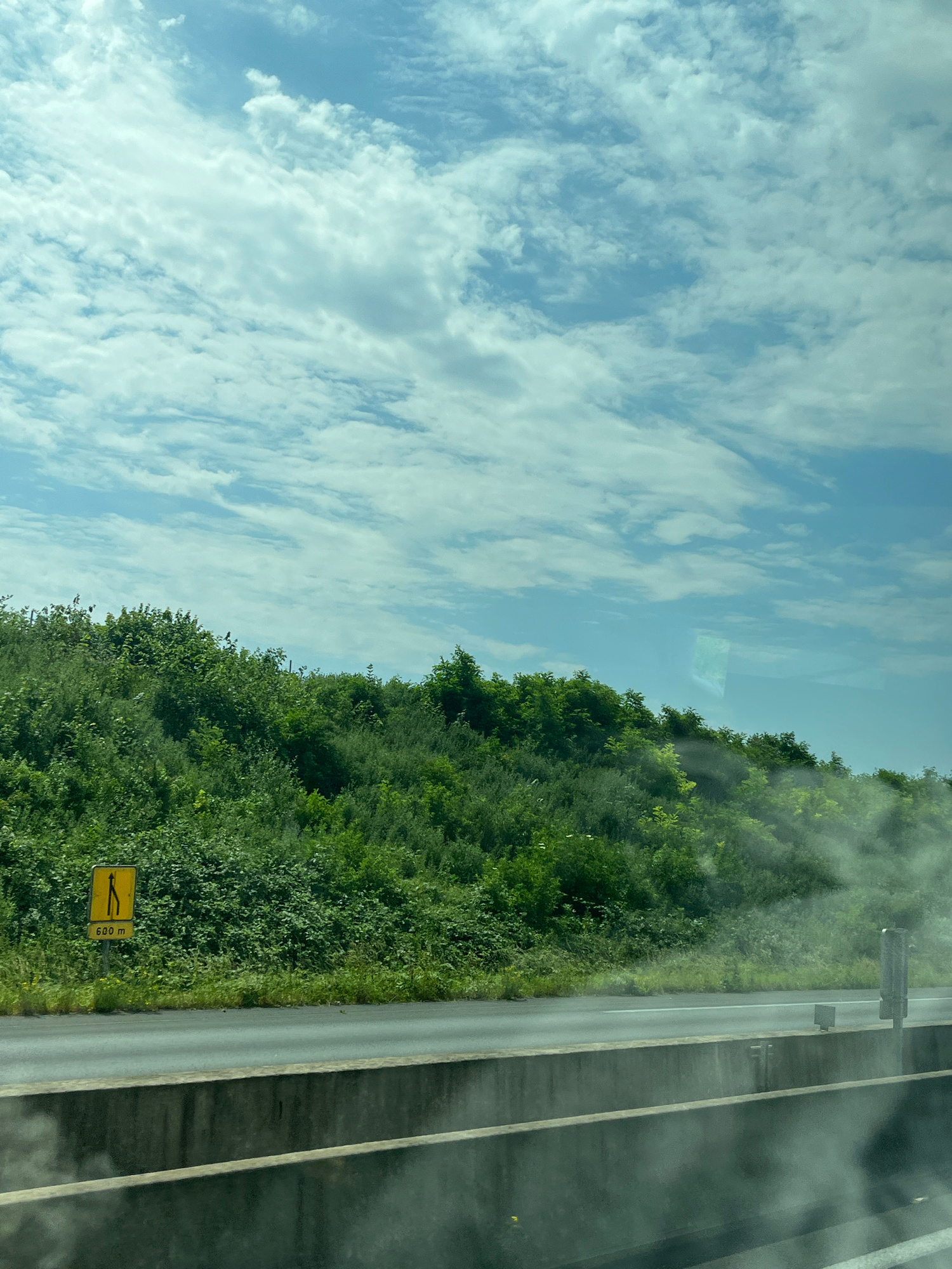 The image captures a view from a vehicle, likely moving along a highway, with a lush green hillside on the right and a bright blue sky with scattered white clouds above. In the foreground, there is a concrete barrier, indicating the perspective is from within a car. A yellow road sign with a black symbol of two diverging paths and the text "600 m" is visible on the left side of the road, suggesting an upcoming split or merge. The environment is predominantly natural, with dense vegetation covering the hillside and the road appearing to be asphalt. There are no visible people or specific urban structures within the frame. The lighting suggests it is daytime, with sunlight illuminating the scene and casting soft shadows. There is a reflection on the surface of the window, obscuring some of the detail. The location of Ramillies, France is not visually ascertainable from the image alone.
