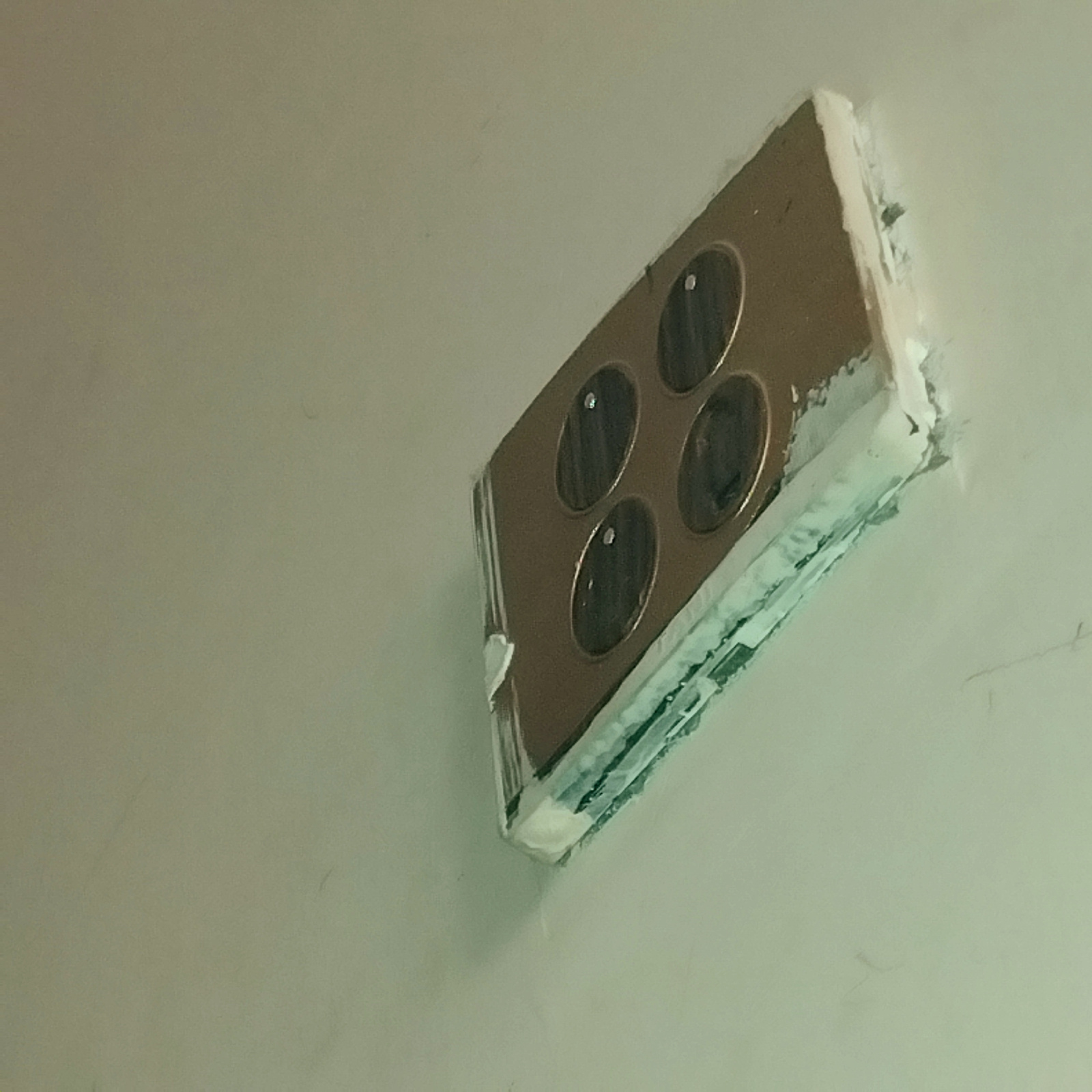 The image shows a close-up, angled view of a metallic-looking object embedded in a light-colored wall. The object is rectangular with rounded corners and has four circular indentations arranged in a 2x2 grid. These indentations appear to be dark and recessed, with some reflective glints within them. The object's edges are slightly rough, and there is some white material, possibly spackle or sealant, around its perimeter, suggesting it has been recently installed or is a part of the wall's construction. The wall itself appears to be a neutral, possibly painted surface, with some subtle texture and faint marks or scratches visible. The lighting in the scene is soft and diffuse, casting minimal shadows. No people, vehicles, or specific landmarks are visible. The overall impression is of a functional component integrated into an interior wall.
