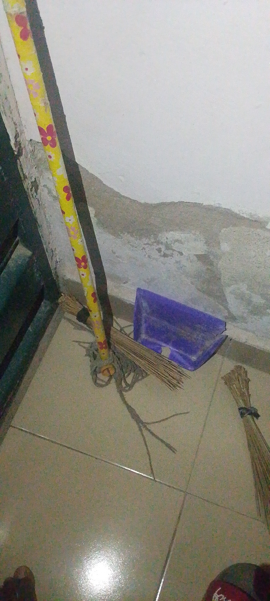 A low-angle, medium shot captures a cleaning setup in a room with tiled flooring and a textured white wall. The focal point is a broom with a yellow handle decorated with red and white flowers. The broom head, made of natural fibers, is resting on the floor. Next to the broom is a purple dustpan. To the right, another broom head, also made of natural fibers and tied with a dark band, lies on the floor. The environment suggests a domestic or utility space. The image does not provide any visual clues to its specific location within Jauro Yinu, Nigeria.