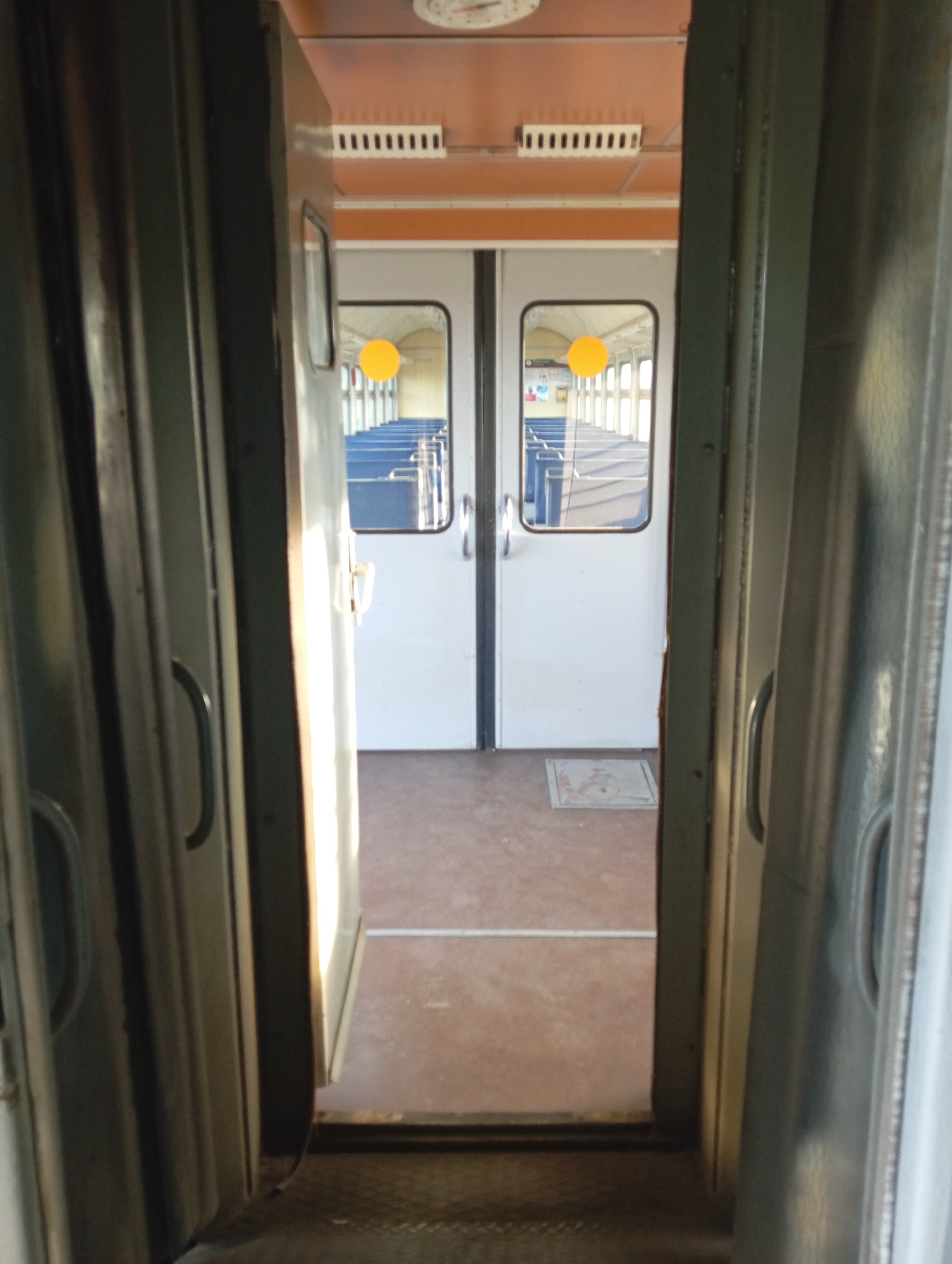 The image shows a view from inside a train carriage looking out through an open doorway. The scene is of the interior of another train carriage. Two doors are visible, each with a rectangular window. Through the windows, rows of blue seats are visible, with white overhead luggage racks and yellow lights reflecting off the windows. One of the windows has a bright yellow circular sticker on it. The walls of the carriage are a light color, and there are some vertical elements that appear to be heating elements or decorative panels on the outer wall. The overall impression is that of a clean and functional train interior. The lighting suggests it is daytime. The location is Artyshta, Russia. There is no text visible in the image.