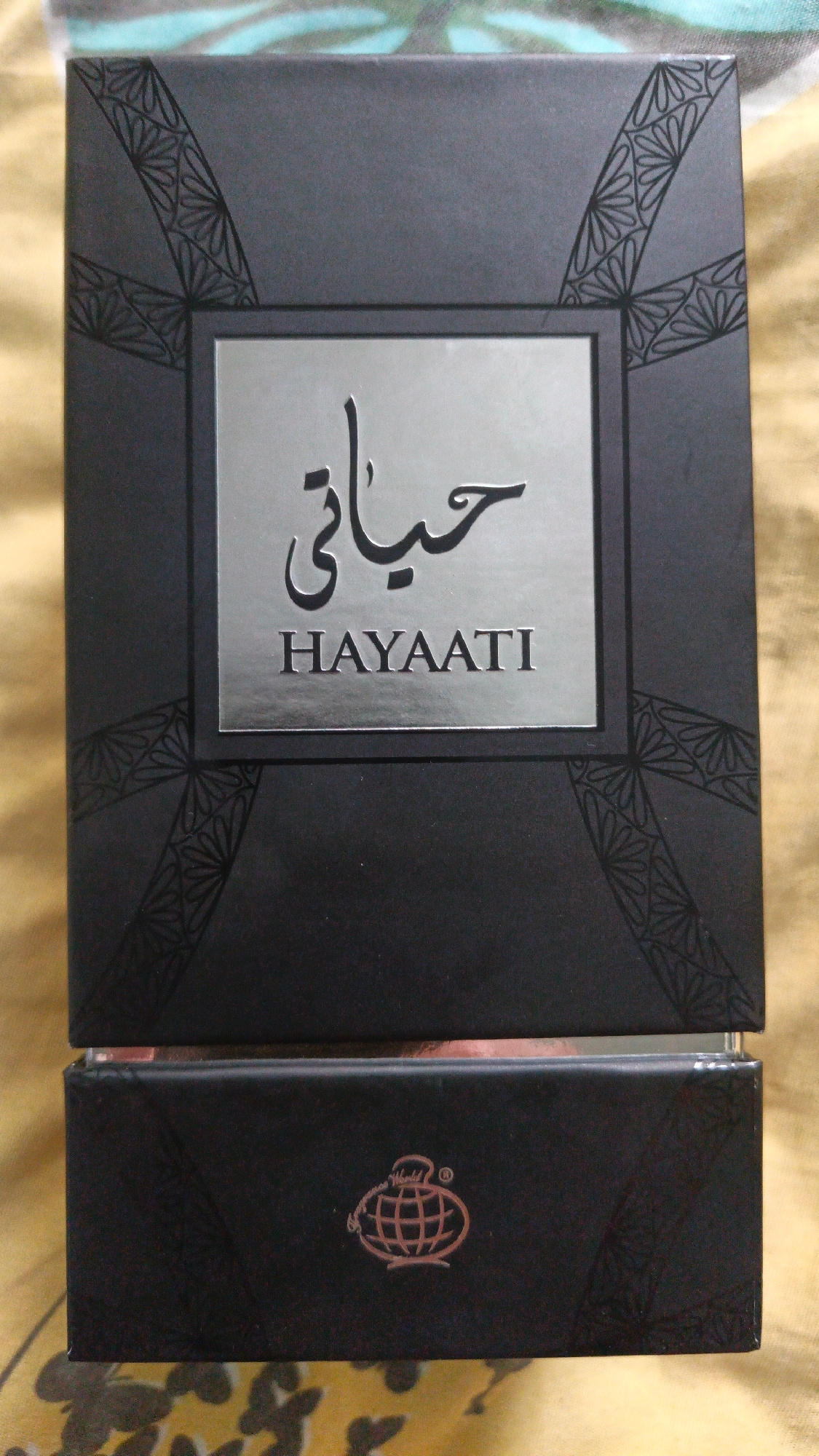 A close-up, slightly overhead shot shows a black rectangular box with metallic silver lettering. The box is oriented vertically and sits on a textured yellow surface. The top portion of the box features a geometric pattern along the top edge and a central metallic silver rectangle containing Arabic script above the English word "HAYAATI." The bottom portion of the box has a reddish-brown hue and displays a metallic copper logo of a globe with a stylized perfume bottle inside. The box appears to be for a fragrance product.