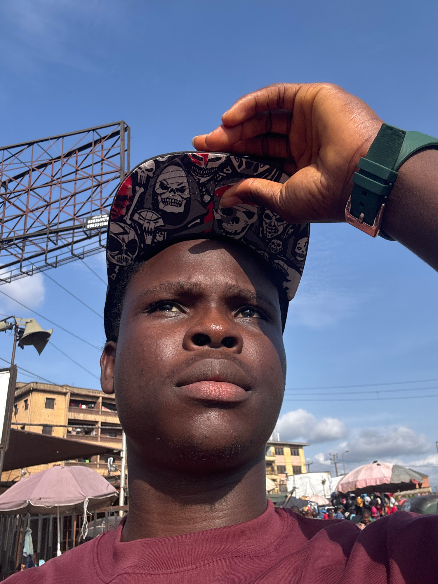 A young Black male is captured in a low-angle, close-up selfie taken outdoors in Onitsha, Nigeria, under a clear blue sky with scattered clouds. He is wearing a maroon t-shirt and a cap adorned with a skull pattern, which he is adjusting with his right hand. A dark green wristwatch with a rose gold buckle is visible on his left wrist.
The background reveals an urban environment. Prominently featured is the skeletal structure of a large billboard. To the left and further back, a multi-story building with balconies and windows is visible. In the mid-ground, market stalls with pink and beige umbrellas are set up, and a variety of goods, possibly clothing, are displayed. The overall scene suggests a bustling street or market area in Onitsha.