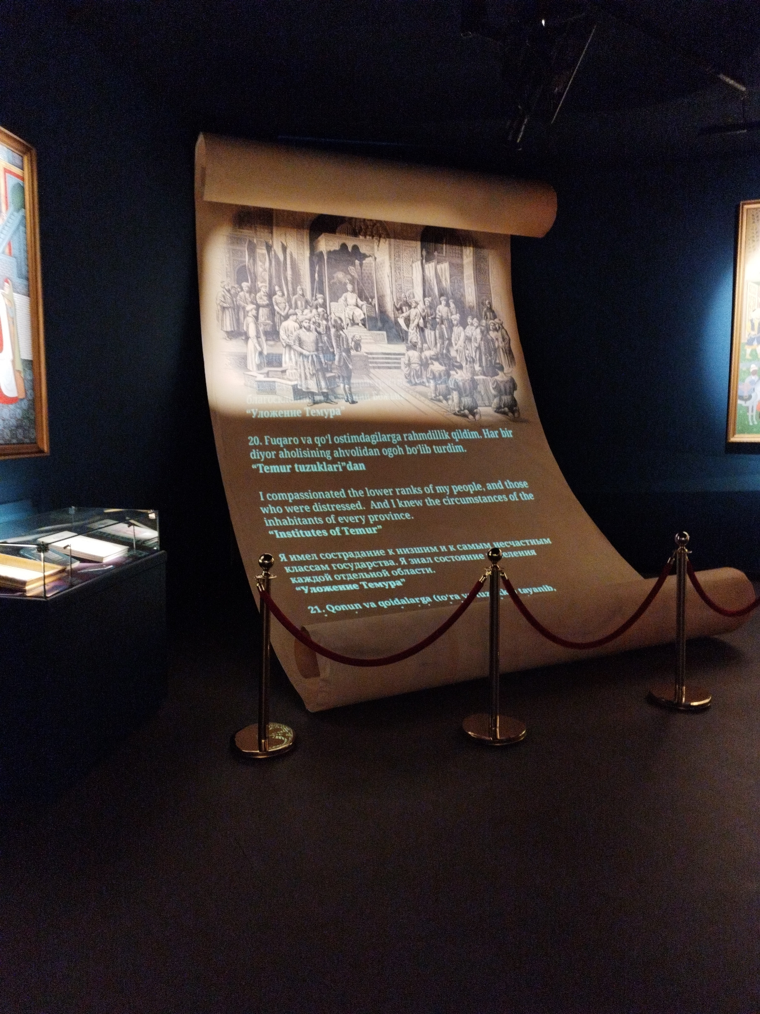 This image depicts an exhibition showcasing a large, unrolled scroll with text and an illustration. The scroll is the central focus, displaying a historical scene of people gathered around a seated figure, likely a ruler, in what appears to be a formal setting with architectural elements. The illustration is rendered in a monochromatic style, suggesting an ancient or historical document.
The scroll is strategically displayed within a museum or exhibition space, evidenced by the velvet ropes and stanchions that cordon off the display. These barriers indicate that the scroll is a valuable artifact being protected from public contact. The surrounding environment is dark, with subtle lighting highlighting the scroll and a portion of a framed artwork on the left side of the image. The artwork appears to be a painting with figures and architectural details, also historical in nature.
Visible text on the scroll is presented in multiple languages. There is a heading in Cyrillic script, "Уложение Темура," which translates to "The Code of Timur." Below this, there is text in what appears to be Uzbek or a related Turkic language, followed by an English translation of a quote, "I compassionated the lower ranks of my people, and those who were distressed. And I knew the circumstances of the inhabitants of every province. 'Institutes of Temur'." Further down, there is more text in Cyrillic, also in Russian, and then more Uzbek. The presence of these multiple languages suggests the exhibition aims to cater to a diverse audience and highlight the historical significance of Timur (Tamerlane) and his legal and administrative principles.
The overall atmosphere is one of historical reverence and education, inviting viewers to engage with the past through the presented artifacts and texts. The scene evokes a sense of solemnity and intellectual curiosity.