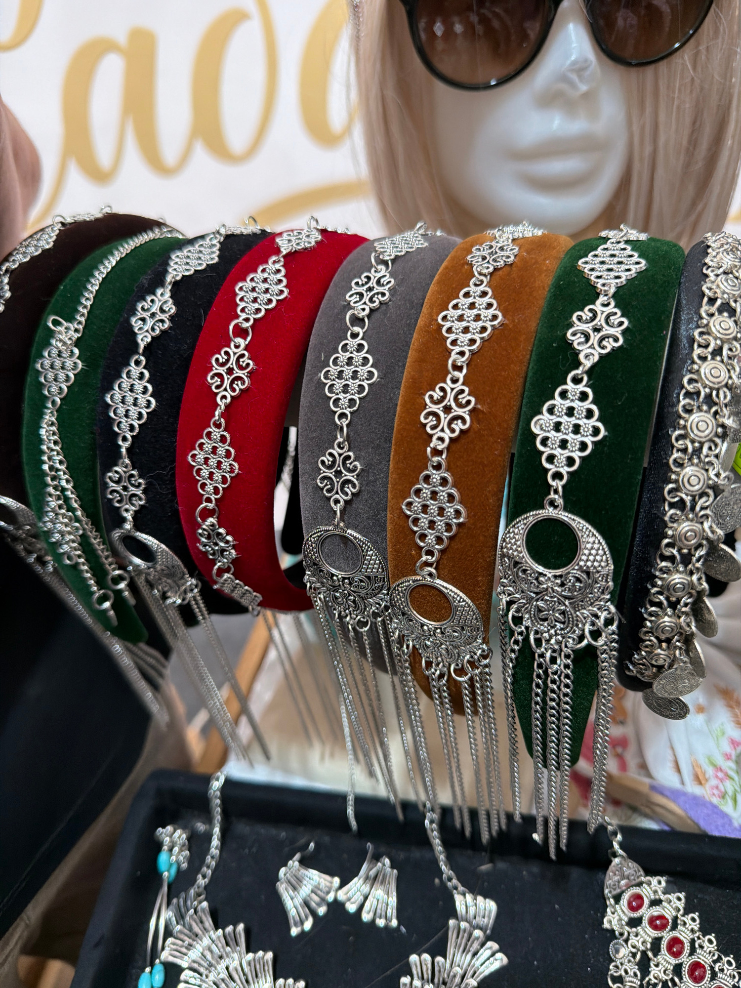 A close-up, overhead shot shows a stack of richly colored, velvet headbands adorned with ornate silver metalwork. The headbands are stacked vertically, with the top one appearing black, followed by green, brown, gray, red, and then another black band. Each headband features a continuous silver chain with decorative filigree and circular medallions. Hanging from the medallions are numerous delicate silver chains, creating a fringe effect. To the left of the headbands, a partial view of a mannequin head with blonde hair and sunglasses is visible. In the background, to the right, a black tray holds more silver jewelry, including what appears to be a necklace with turquoise-colored beads. The overall impression is of a display of handcrafted accessories, likely in a retail or market setting. The lighting is bright, suggesting an indoor environment or a shaded outdoor area. No visible text is present in the image.