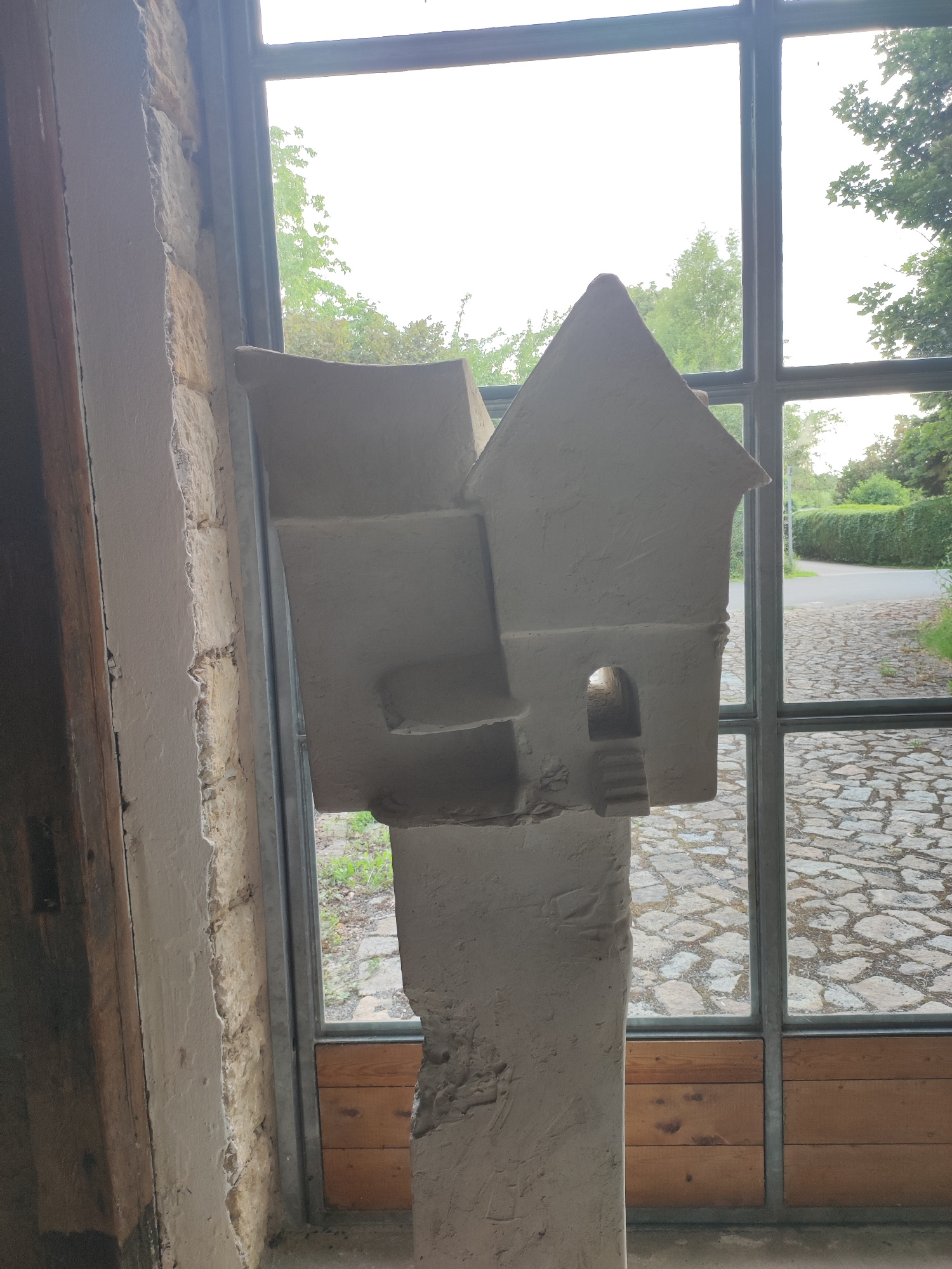A light-colored, abstract sculpture resembling a multi-level building with an arched entryway and stairs is positioned in the foreground. The sculpture is made of a rough, plaster-like material and stands on a similarly textured pedestal. Behind the sculpture, a large window with dark metal framing offers a view of an outdoor scene. The exterior features a cobblestone path leading to a road lined with hedges and trees. The sky is bright and overcast. The architectural style of the window and the stonework visible on the left suggest a historical building. This scene is located at the former "Vereinigte Sächsische Hütten und Stahlwerke" (United Saxon Foundries and Steelworks) in Dresden, Germany, which has been converted into a cultural center known as the Kulturpalast Dresden.