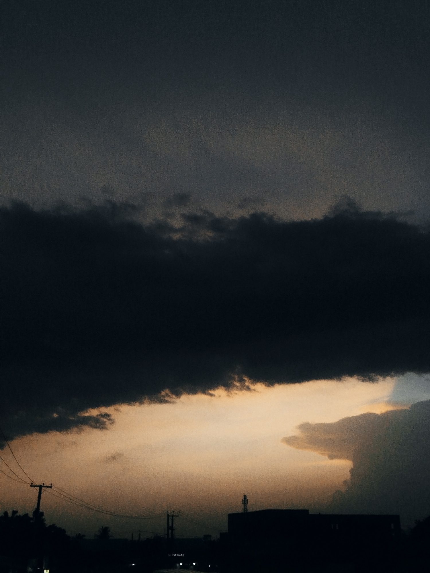 A dramatic sky fills the frame, dominated by a large, dark cloud formation stretching horizontally across the upper half of the image. Beneath this dark mass, a band of warm, orange-yellow light emanates from the horizon, illuminating thinner clouds and the silhouette of structures in the foreground.
The foreground features a dark, silhouetted cityscape. On the left, a prominent utility pole with wires is visible against the lighter sky. Further to the right, other utility poles and silhouetted structures, possibly buildings, are discernible. One building on the right has a small antenna or structure on its roof. The overall impression is one of an impending storm or the aftermath of sunset in an urban environment. The location is identified as Narayi, Nigeria, suggesting an African cityscape. There are no visible people or animals in the image.