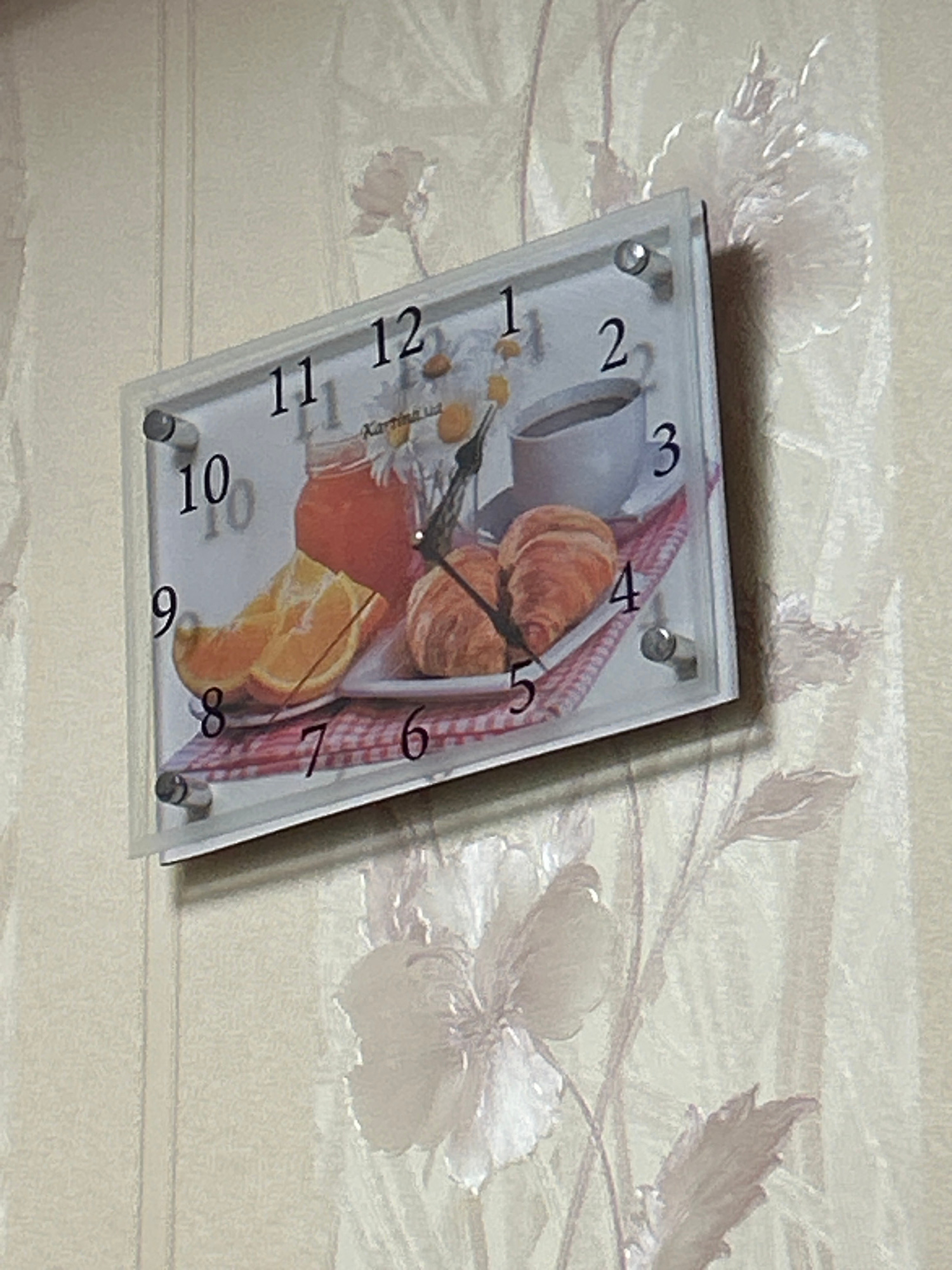 The image shows a close-up, slightly angled view of a rectangular wall clock with a white background and black numbers. The clock face features an illustration of breakfast items, including orange slices, a jar of jam, two croissants, and a cup of coffee. The clock hands are black, with the hour hand pointing towards the "10" and the minute hand pointing towards the "2". The clock is mounted on a beige wallpaper with a subtle floral pattern. The overall scene is static and appears to be an indoor shot. The image does not contain any people or provide any information about the location in Ivanivka, Ukraine.
