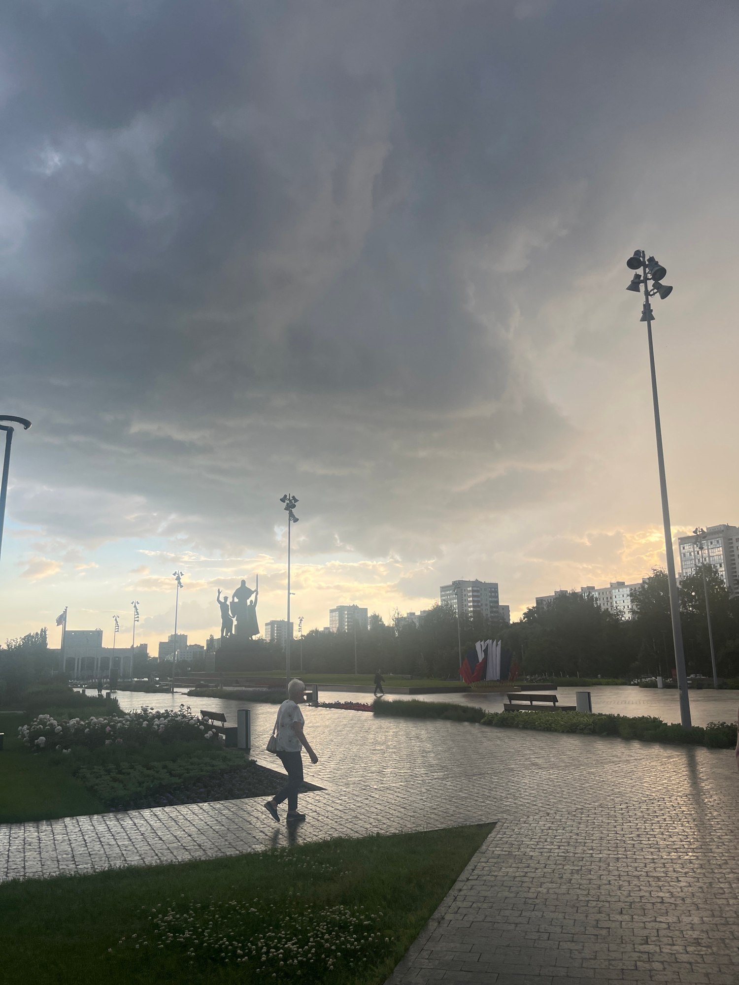 The image shows a paved walkway in a park-like setting, likely in the city of Perm, Russia, given the distinctive monument visible in the background. The walkway is made of brick pavers, wet from recent rain, reflecting the overcast sky. To the left of the walkway, there is a manicured lawn with scattered white flowers and a raised garden bed with green foliage and some flowering plants. A woman is walking along the walkway from left to right. She is wearing a light-colored blouse and dark pants, and carrying a light-colored purse over her shoulder. In the middle ground, there is a large, dark monument depicting figures. Beyond the monument, several tall, modern buildings are visible, silhouetted against a sky with dramatic clouds that are breaking to reveal patches of sunlight. Several lampposts are also visible, including one prominently to the right of the frame. In the far background, more buildings and trees can be seen. The overall impression is a somewhat somber atmosphere due to the cloudy sky, but with hints of brightness breaking through.