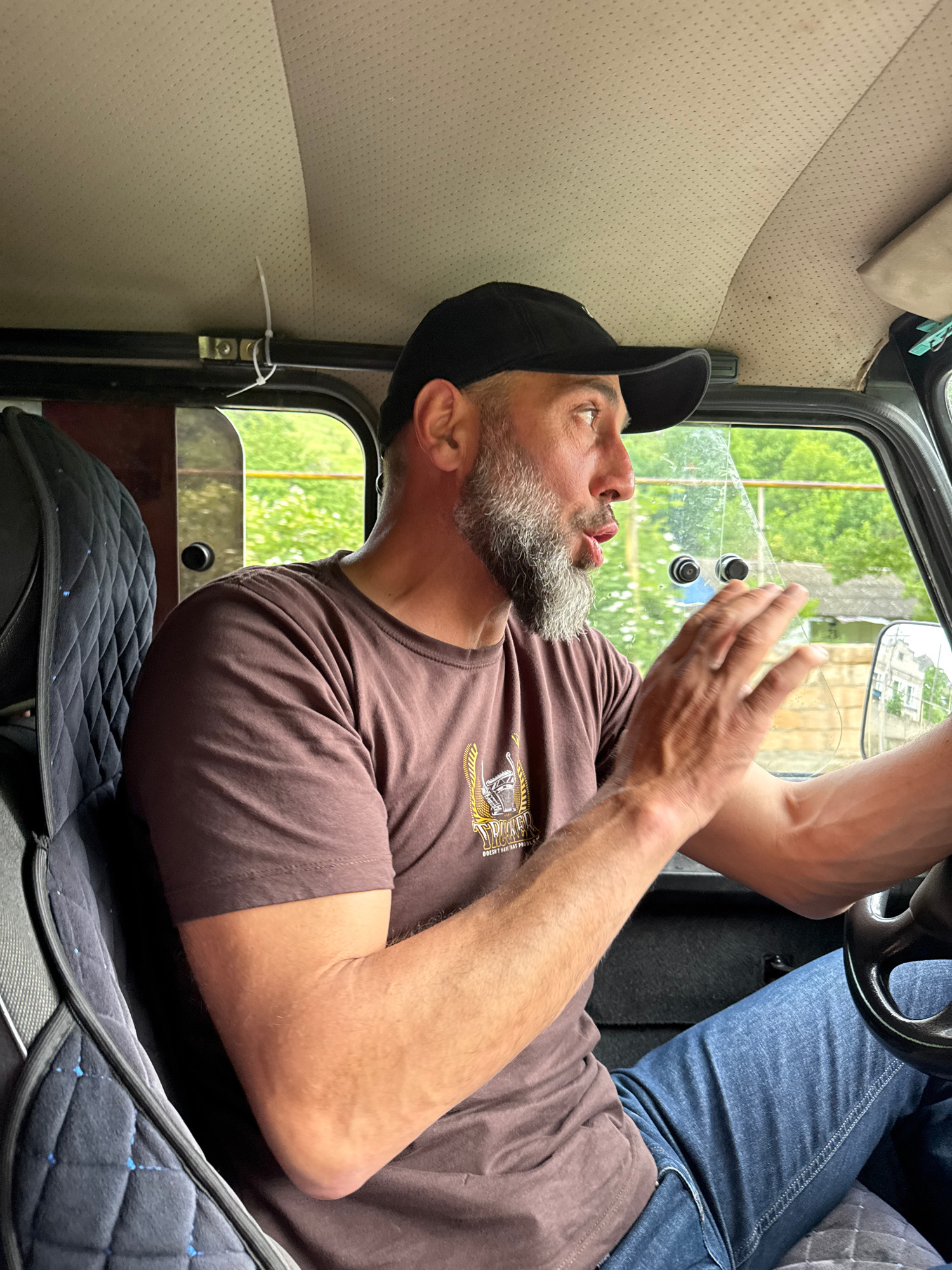 A medium shot shows a bearded man with a graying beard and a black baseball cap driving a vehicle. The man is wearing a brown t-shirt with a graphic and blue jeans. He is looking to his right, with his mouth open and his right hand raised as if speaking or gesturing. The interior of the vehicle is visible, including the beige headliner with a cable tie securing something to it, a quilted dark blue seat, and the steering wheel. Outside the window, green foliage and a building are visible. The image appears to be taken in Каменномостский, Russia.