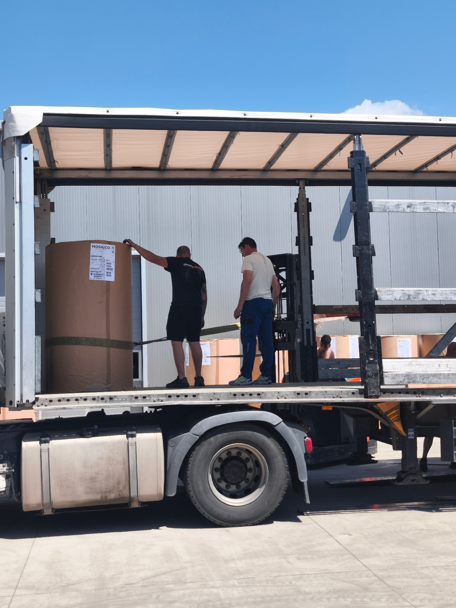 Two men are loading a large cylindrical object wrapped in brown paper onto the back of a truck. The truck's trailer is open, revealing the interior where more boxes are stacked. One man, wearing a black t-shirt and shorts, is standing next to the cylinder, touching it with his right hand. The other man, in a light-colored t-shirt and blue pants, is standing further back, near a piece of machinery that appears to be a forklift. The scene takes place outdoors on a bright, sunny day, with a clear blue sky overhead. The background features a light-colored, ribbed building. The truck's tires and fuel tank are visible in the foreground.