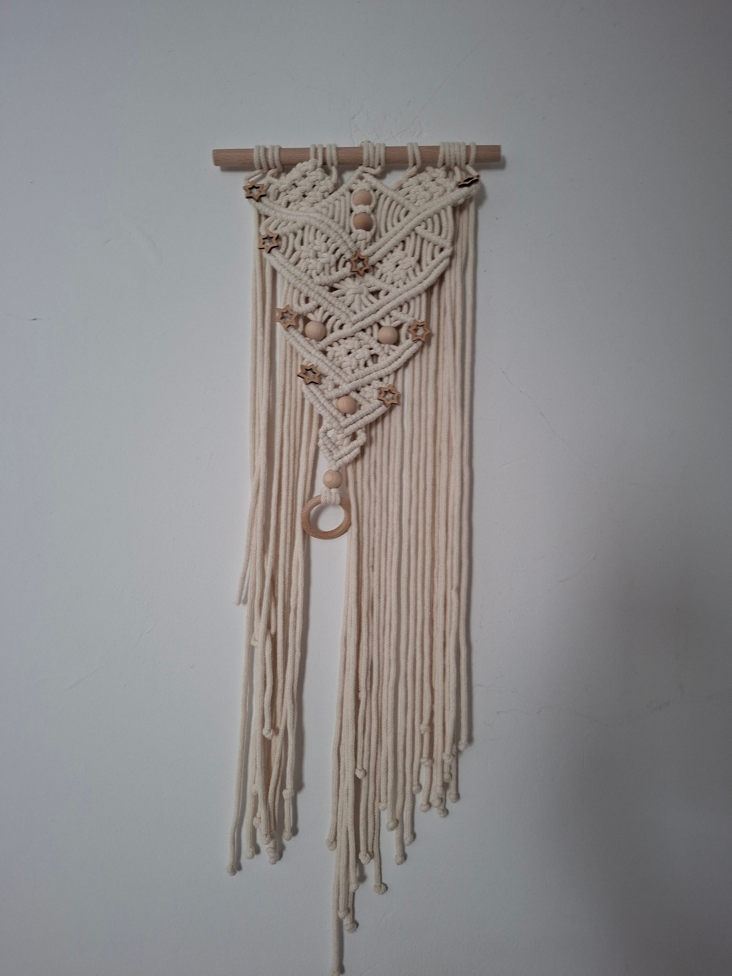 A macrame wall hanging is displayed on a plain, light-colored wall. The wall hanging is made of thick, cream-colored cord and is suspended from a natural wood dowel. The design features intricate knotting that forms a diamond shape in the upper portion, embellished with several small, star-shaped wooden beads and round wooden beads. Below the diamond shape, the cord hangs in a fringe, with a single wooden ring as a pendant near the bottom. The wall appears to be a standard interior wall with minor imperfections. No people or other objects are visible in the frame. The lighting is soft, suggesting an indoor environment. The location is unknown.