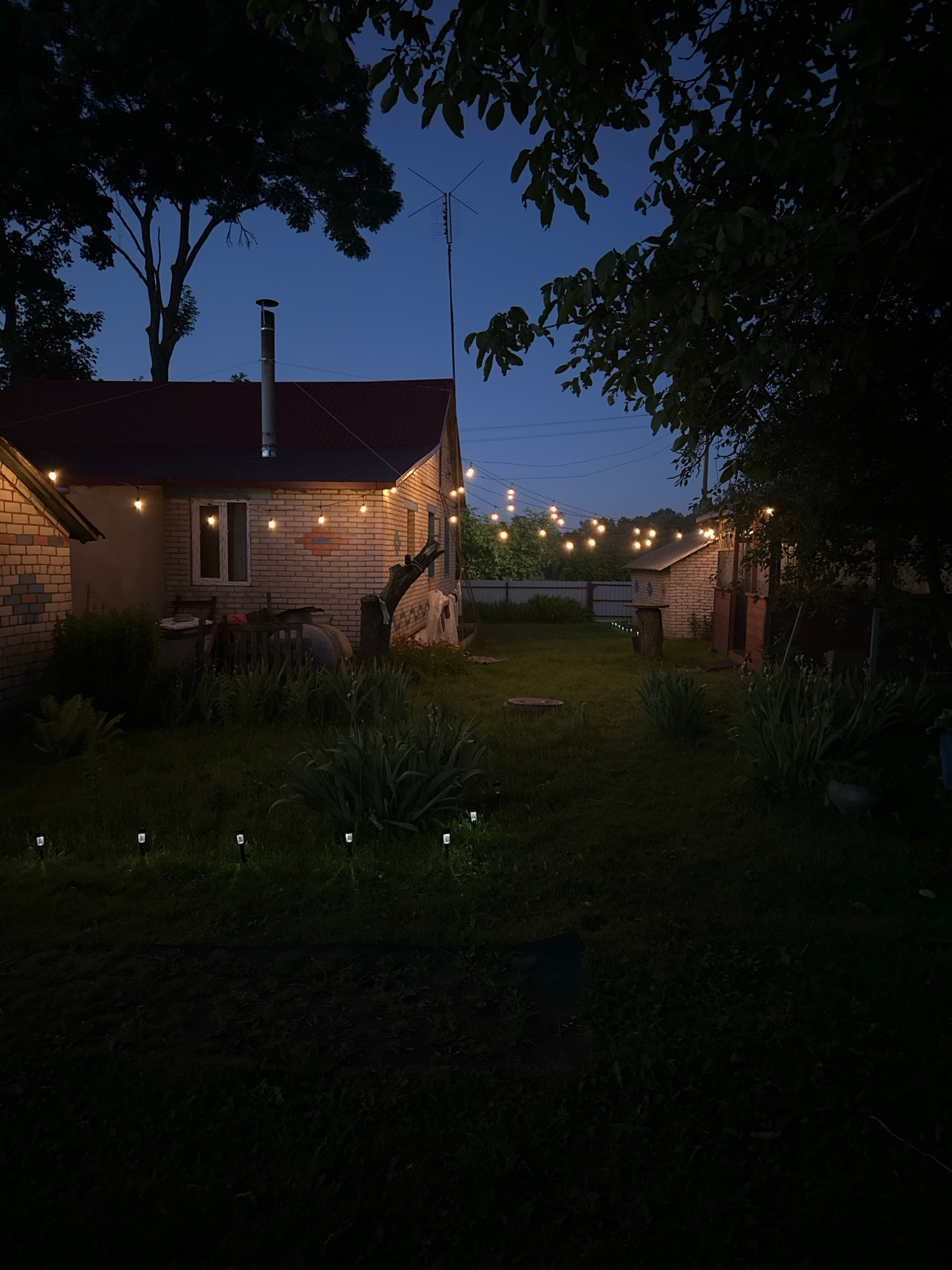 The image depicts a backyard scene at dusk, illuminated by string lights. The foreground is dominated by dark foliage, with a large tree on the left silhouetted against a deep blue sky. A residential building, constructed from light-colored brick, is visible on the left side of the frame. It has a dark, pitched roof and several windows, one of which is lit from within. A metal chimney extends from the roof.
A string of warm, glowing Edison-style bulbs is suspended across the yard, casting a festive light. These lights stretch from the left side of the image, passing by the building, and extending into the background. In the background, more of the brick building is visible, along with what appears to be a fence and some vegetation.
The yard itself is covered in green grass, with several smaller, square-shaped lights planted along the edge of the lawn, creating a subtle pathway. The overall mood is serene and inviting, suggesting an evening gathering or a quiet moment outdoors. The time of day is clearly indicated by the twilight sky and the artificial lighting. No text is visible in the image.