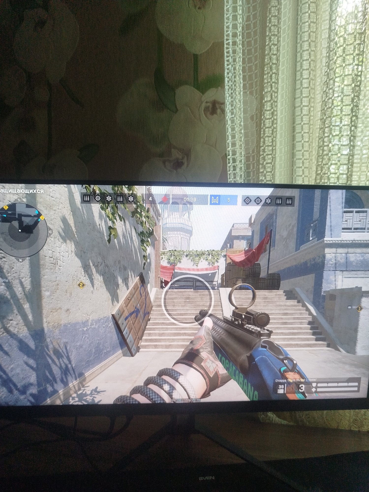 The image displays a screenshot from a video game, depicting a first-person perspective. The player is holding a rifle with a scope, aiming down a set of stairs. The environment appears to be an urban setting with buildings characterized by a mix of concrete walls and blue painted sections. Green foliage cascades down a wall on the left side of the screen. A red banner is visible in the distance, tied to a pole. The game interface shows a timer at "01:39", a player count of "5", and icons representing ammunition and possibly other in-game elements. A minimap is present in the upper left corner. The bottom right of the screen shows the weapon's name "Chiappa Triple Threat" and the player's current ammo count of "39" with "3" in reserve. The overall scene is a typical representation of a combat scenario within a video game. There is no visible information or context within the image that places this scene in the city of Rudny, Kazakhstan.