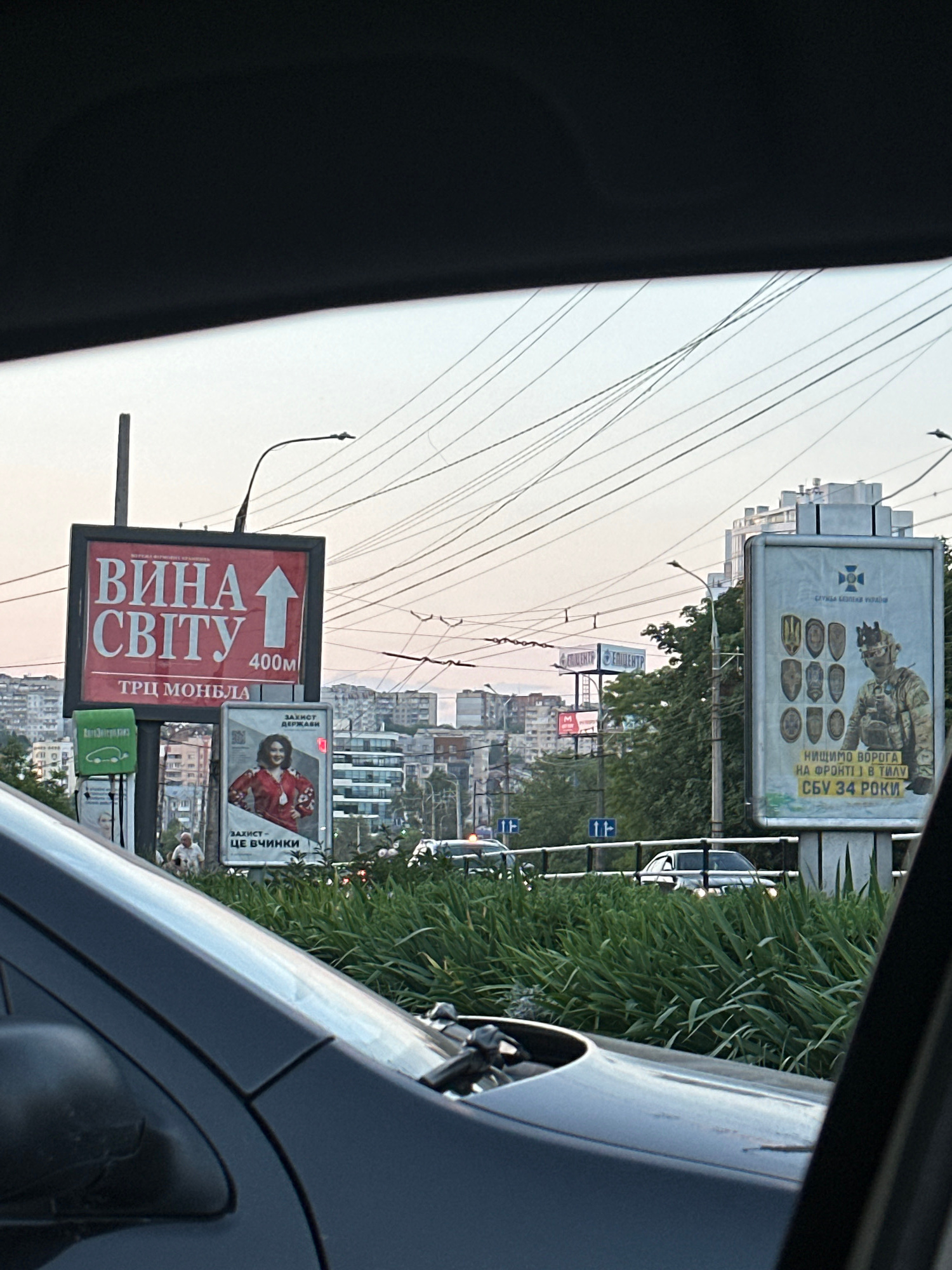 The image captures a street scene in Khmelnytskyi, Ukraine, as viewed from inside a vehicle. In the foreground, the dark gray hood and windshield of a car are visible, obscuring the lower portion of the frame. Beyond the car, a vibrant green hedge lines a street.
Dominating the middle ground are several billboards. The most prominent is a large, red billboard with white Ukrainian text that reads "ВИНА СВІТУ" (Wines of the World) and an upward-pointing arrow indicating "400м". Below this, it says "ТРЦ МОНБЛА" (TRC Monbla). To the right of this billboard, a smaller billboard features a woman in a traditional embroidered shirt, with text below that translates to "PROTECTION - THIS IS THE WORK OF WOMEN".
Further to the right, another large billboard displays an image of a soldier in military gear, surrounded by shields, with Ukrainian text that reads "WE ARE STRIKING THE ENEMY ON THE FRONT LINE AND IN THE REAR THE SBU IS 34 YEARS OLD". This billboard is associated with the Security Service of Ukraine (СЛУЖБА БЕЗПЕКИ УКРАЇНИ).
In the background, multi-story apartment buildings and other structures of a city are visible under a pale sky with numerous overhead wires. The lighting suggests it is either early morning or late evening. A few pedestrians and vehicles are present on the street, but they are not the focus of the image. The overall impression is of a city environment with advertising and everyday life unfolding.