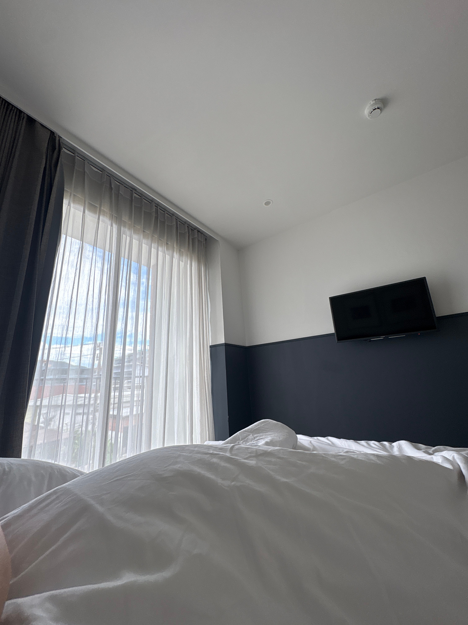 The image shows a hotel room in Chalong, Thailand. The perspective is from a bed, looking towards a window and a wall. The bed is covered with white linens and appears to be rumpled, suggesting it has been slept in. Sheer white curtains hang over the window, which looks out onto a cityscape with buildings and the sky. A darker curtain is partially visible on the left side of the window. On the wall, a flat-screen television is mounted above a dark blue section of the wall, which contrasts with the white upper portion. A smoke detector is visible on the white ceiling. The room is brightly lit, likely by natural light from the window.