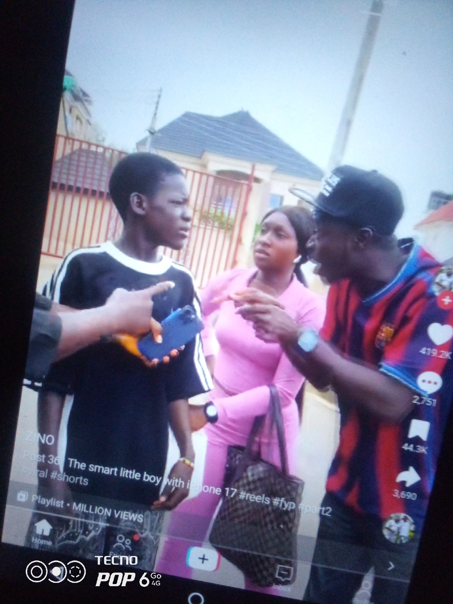 The image is a slightly distorted, angled view captured from a screen, likely a smartphone, displaying a social media video. In the foreground, three individuals are visible. On the left, a young person wearing a black t-shirt with white stripes on the sleeves and collar is holding a blue smartphone. Their left arm is extended, seemingly interacting with the phone. To their right, a woman in a pink long-sleeved top is looking towards the smartphone. To the far right, a person wearing a black baseball cap and a red and blue striped t-shirt is gesturing with their right hand. A dark patterned tote bag is slung over the woman's shoulder. The background shows a residential area with a beige building and a red metal fence. The sky is overcast. Text overlaid on the bottom of the screen indicates a post title: "The smart little boy with iphone 17 #reels #fyp #part2", and mentions "MILLION VIEWS." Social media interaction icons like a heart, comment bubble, and share arrow are also visible on the right side. The presence of the text "Howayi, Nigeria" is not explicitly visible in the image itself, but is provided as contextual information for the location.