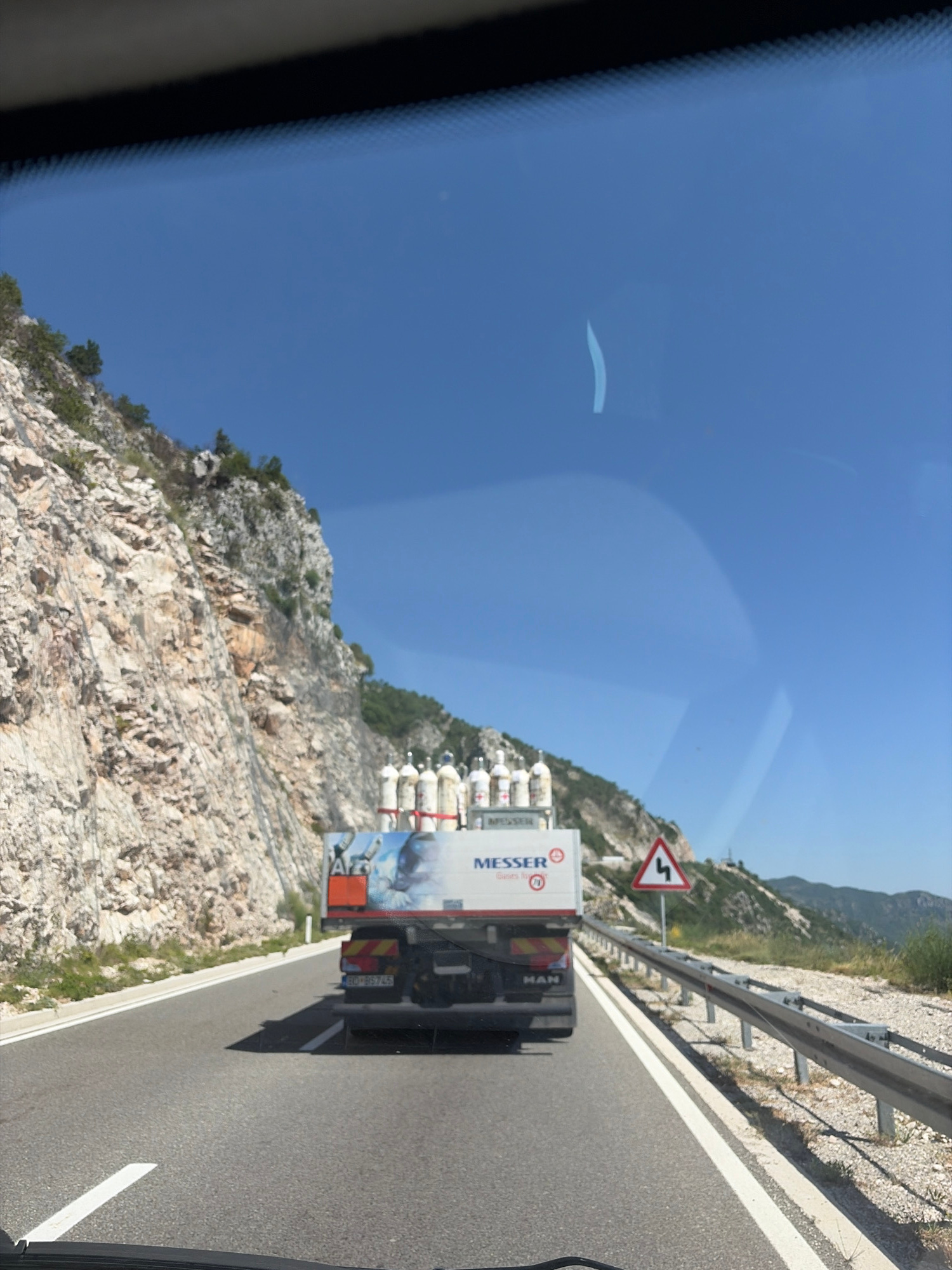This media file depicts a view from a vehicle on a sunny day, likely late morning or early afternoon, given the bright sunlight and clear blue sky. The setting appears to be a mountainous coastal region, possibly along a highway in Brajici, Montenegro, as suggested by the terrain and road features.
The primary objects in the scene are a white truck with the "MESSER" logo and several white cylindrical tanks loaded onto it, suggesting the transport of industrial gases. The truck is driving on a paved road with a white lane marker. To the left of the road, a steep, rocky mountainside with sparse vegetation is visible. A red and white triangular road sign with a symbol indicating a sharp double bend or zigzag road is mounted on a post near the mountainside.
The perspective is from inside a vehicle, indicated by the reflection on the glass, which shows a portion of the dashboard or windshield. The orientation of the image is somewhat tilted, making it appear as if the camera is leaning to the side. There is no discernible emotion conveyed in the static image. The overall impression is a snapshot of travel through a scenic, rugged landscape. Visible text includes the "MESSER" logo on the truck and "MAN" on the back of the truck cab. There is also a license plate on the truck with the characters "BD-BS745".