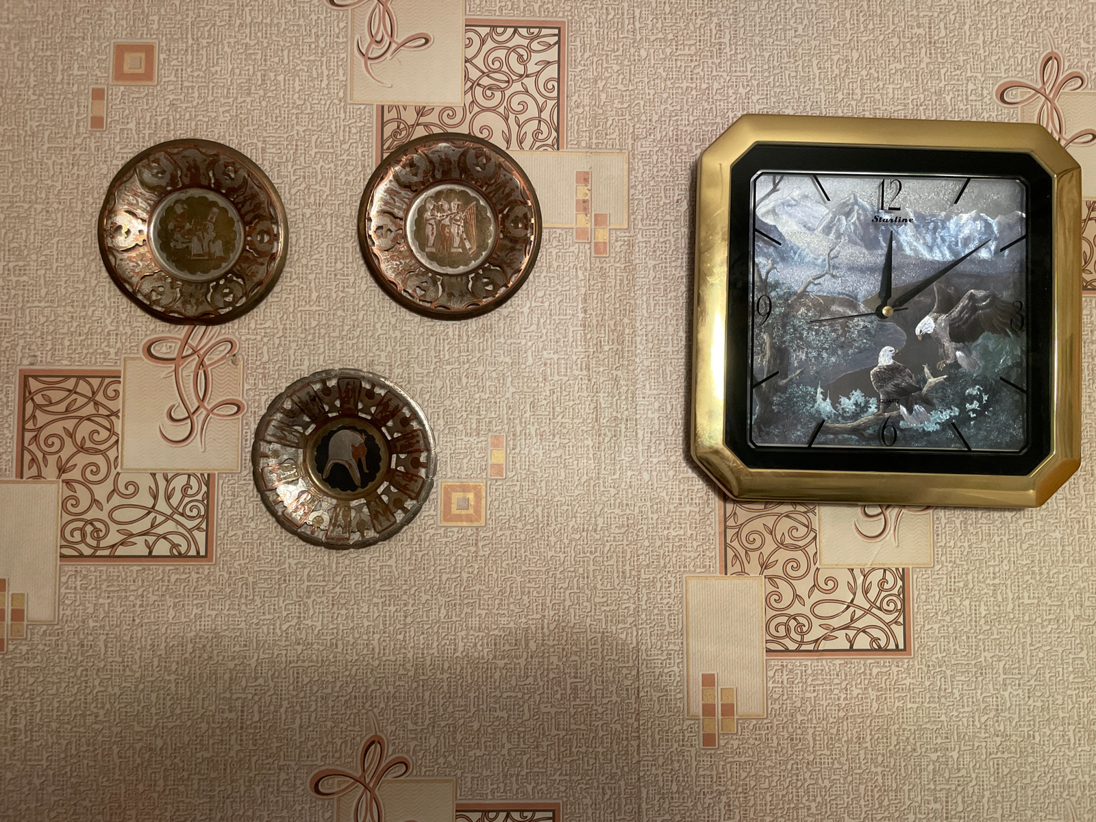 The image shows a wall with wallpaper and decorative elements, including three small decorative plates and a clock. The wallpaper has a textured pattern in beige and cream tones, with some areas featuring ornamental designs in pink and brown.
The three decorative plates are arranged in a vertical line on the left side of the wall. The top plate has a circular design with a scene featuring figures. The middle plate also has a circular design with figures and musical instruments. The bottom plate is circular and appears to have a scene with an animal.
To the right of the plates is a square clock with a gold-colored frame. The clock face displays a scenic image of mountains and eagles. The time shown on the clock is approximately 10:50. The clock has a black bezel and a white face with black hour and minute hands, and a thinner second hand. The brand name "Starline" is visible on the clock face.
The background is the textured wallpaper, which has a repeating geometric and abstract pattern. There are also some decorative square and rectangular elements on the wallpaper, some with swirling patterns and some with a plain texture.
The lighting in the image suggests an indoor environment, likely a room in a home. There are no people or animals visible in the scene other than what is depicted on the plates and the clock. The image does not provide any information about its location in Moscow, Russia.
