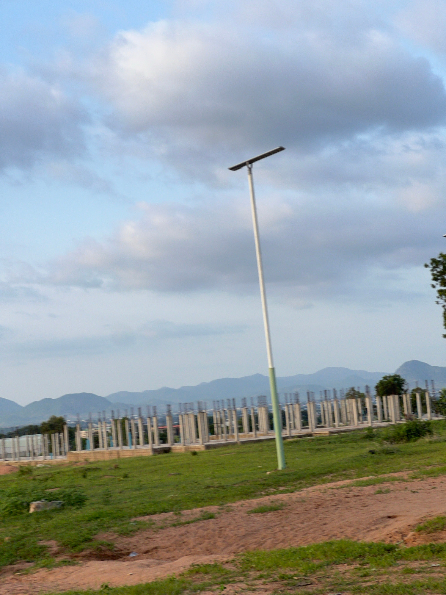 A vertical, long-shot, eye-level photograph captures a light pole and a construction site under a cloudy sky. The light pole, positioned slightly off-center to the right, is a slender, white cylinder with a light green base. It extends from the bottom of the frame towards the top, with a horizontal, rectangular light fixture attached near its apex. The sky above is filled with a mix of white and gray clouds, indicating an overcast or partly cloudy day.
In the middle ground, a construction site is visible. It features numerous concrete pillars arranged in rows, suggesting the foundation or framework of a building in progress. These pillars are situated on a slightly elevated area. Green grass covers the ground around the construction site, with patches of brown, sandy soil in the foreground. In the background, a range of hazy, blue-hued mountains forms a scenic backdrop. The overall environment appears to be an outdoor setting, possibly on the outskirts of a developing area. The city of Jauro Yinu, Nigeria, is the stated location. There are no people visible in the image.