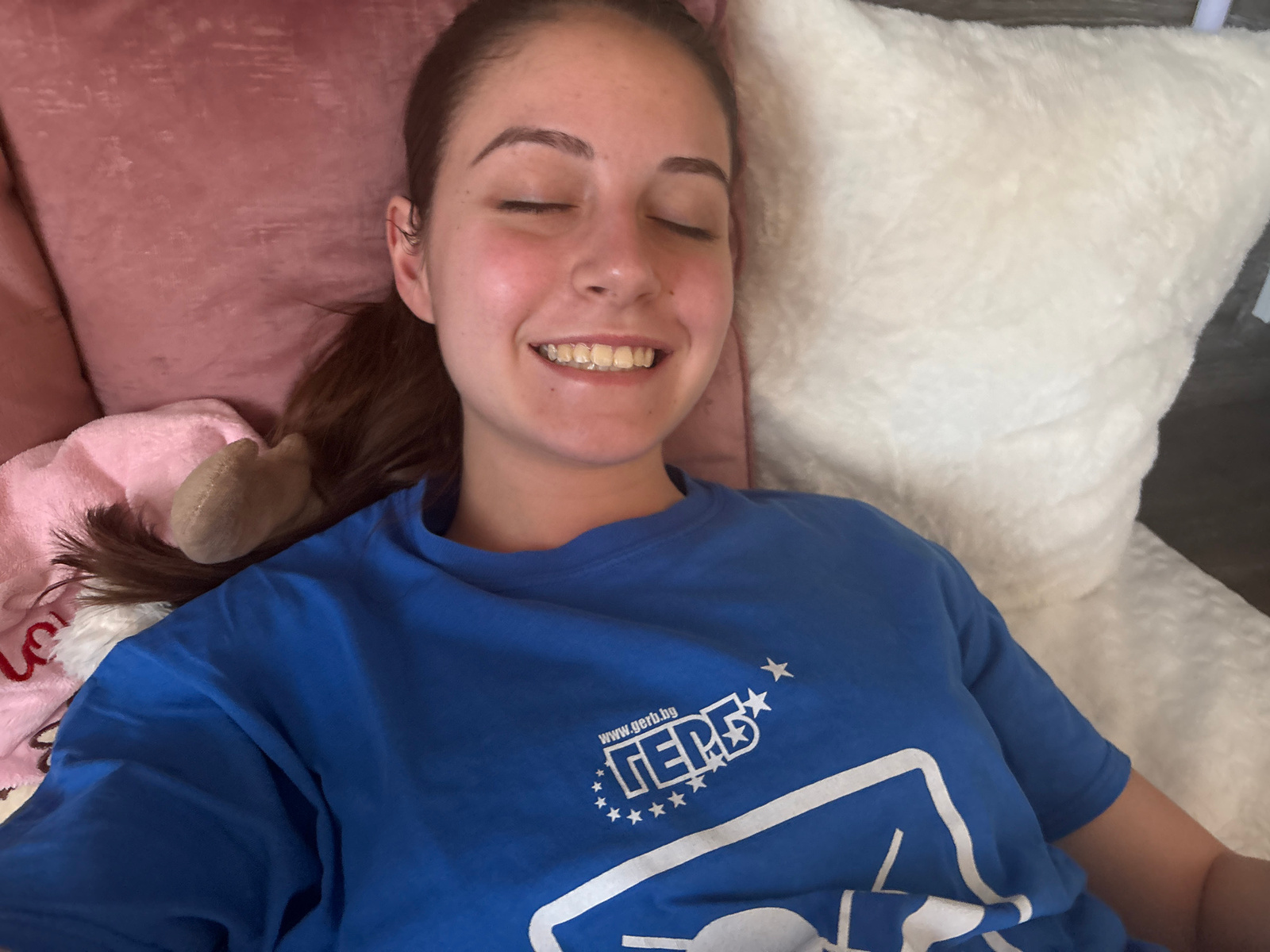 A young woman with her eyes closed and a smile on her face lies on a bed with pink and white pillows, wearing a blue t-shirt with a white logo. The logo features the word "ГЕРБ" (GERB), the name of a political party in Bulgaria, along with a website address and stars. The background includes a glimpse of what appears to be a dark wooden floor and a wall. The presence of the GERB logo strongly suggests the location is in Bulgaria, and given the context provided, it is specifically in the city of Bulgarovo.