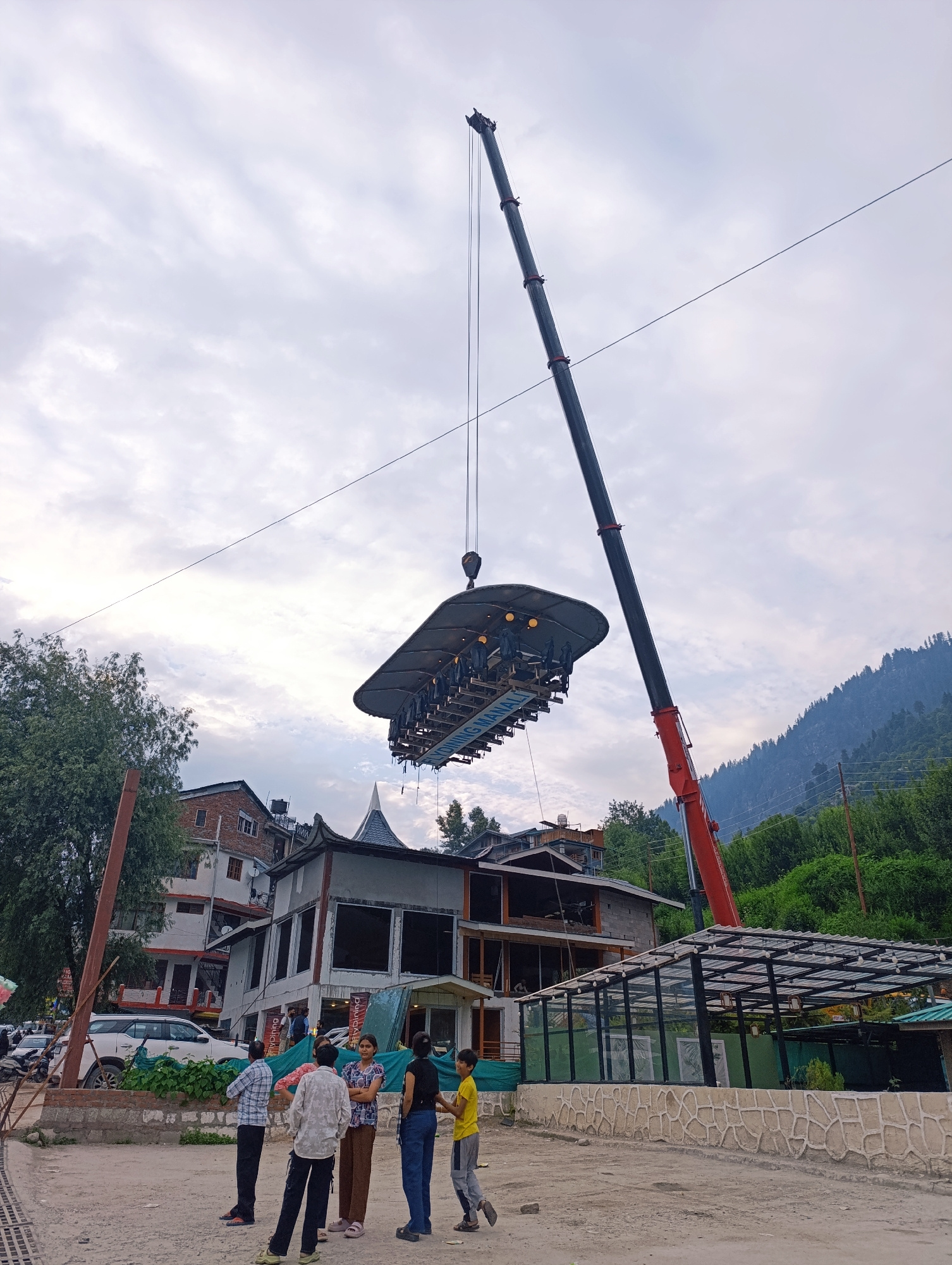 A crane is lifting a unique dining setup, which appears to be a suspended restaurant table with chairs, into the air. The setup is circular with a canopy overhead and lights illuminating the dining area. Several people are seated around the table, looking down or observing the surroundings.
In the foreground, a group of five individuals, including adults and children, stand on a paved area. One person in the foreground is wearing a plaid shirt and another in a white shirt, both looking towards the suspended dining area. A child in a yellow shirt stands to the right of the group, looking to the left.
The scene is set in a town with multi-story buildings and a lush green mountainside in the background. The architecture of the buildings suggests a semi-urban or tourist area. A white SUV is parked on the left side of the image, and some motorcycles are visible further back. The sky is overcast with clouds, indicating a possibly cloudy or humid day. The overall atmosphere appears to be one of curiosity and anticipation, perhaps watching the setup of this unusual dining experience. Text visible on the suspended platform reads "FLY DINING MANALI".
