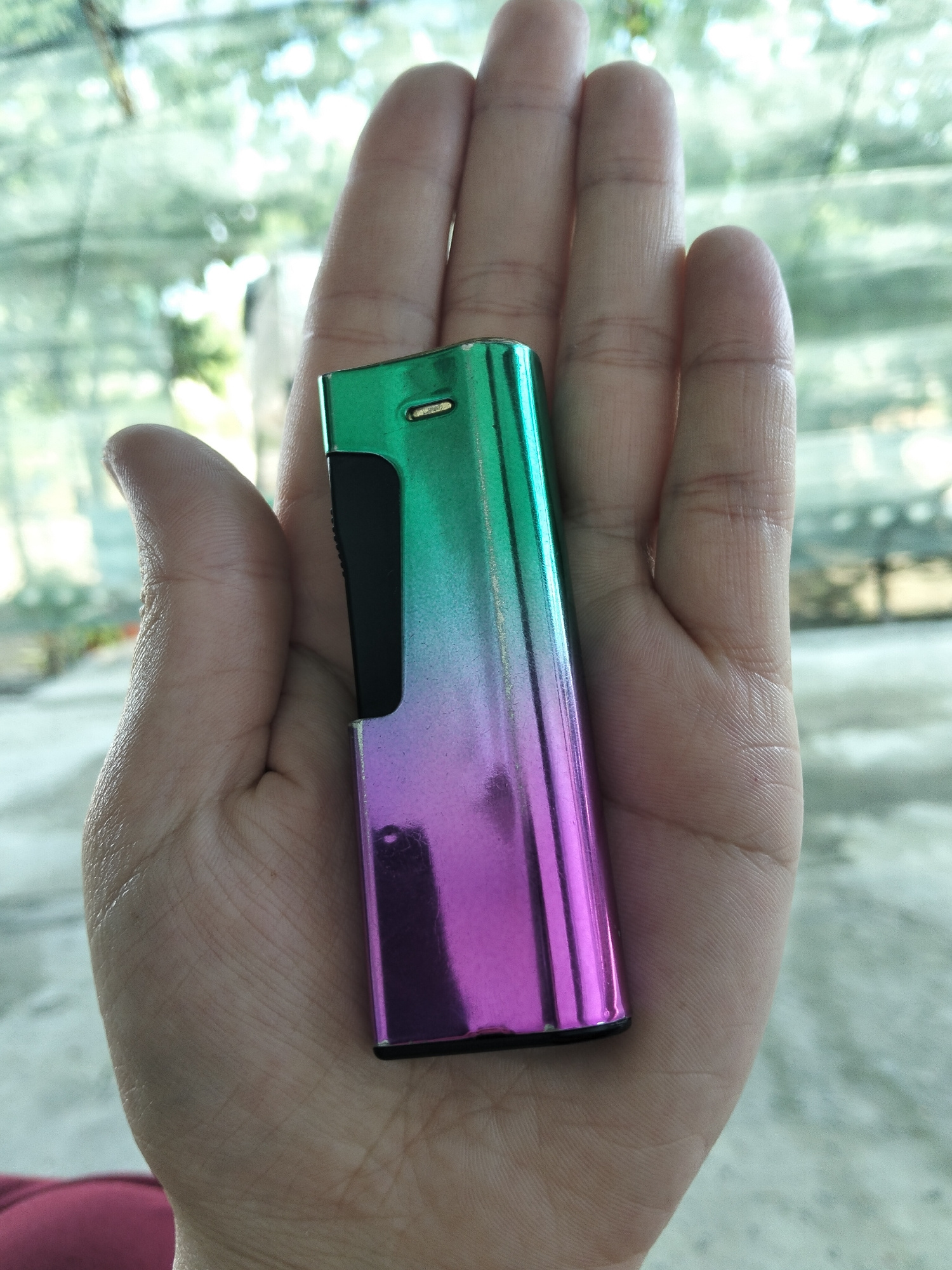 A hand holds a metallic, rectangular object that transitions in color from green at the top to purple at the bottom. The object appears to be a lighter. The hand is resting palm-up, with the object placed in the center of the palm. The background is blurred but suggests an outdoor setting with foliage and possibly a fence or netting. The lighting indicates daytime.